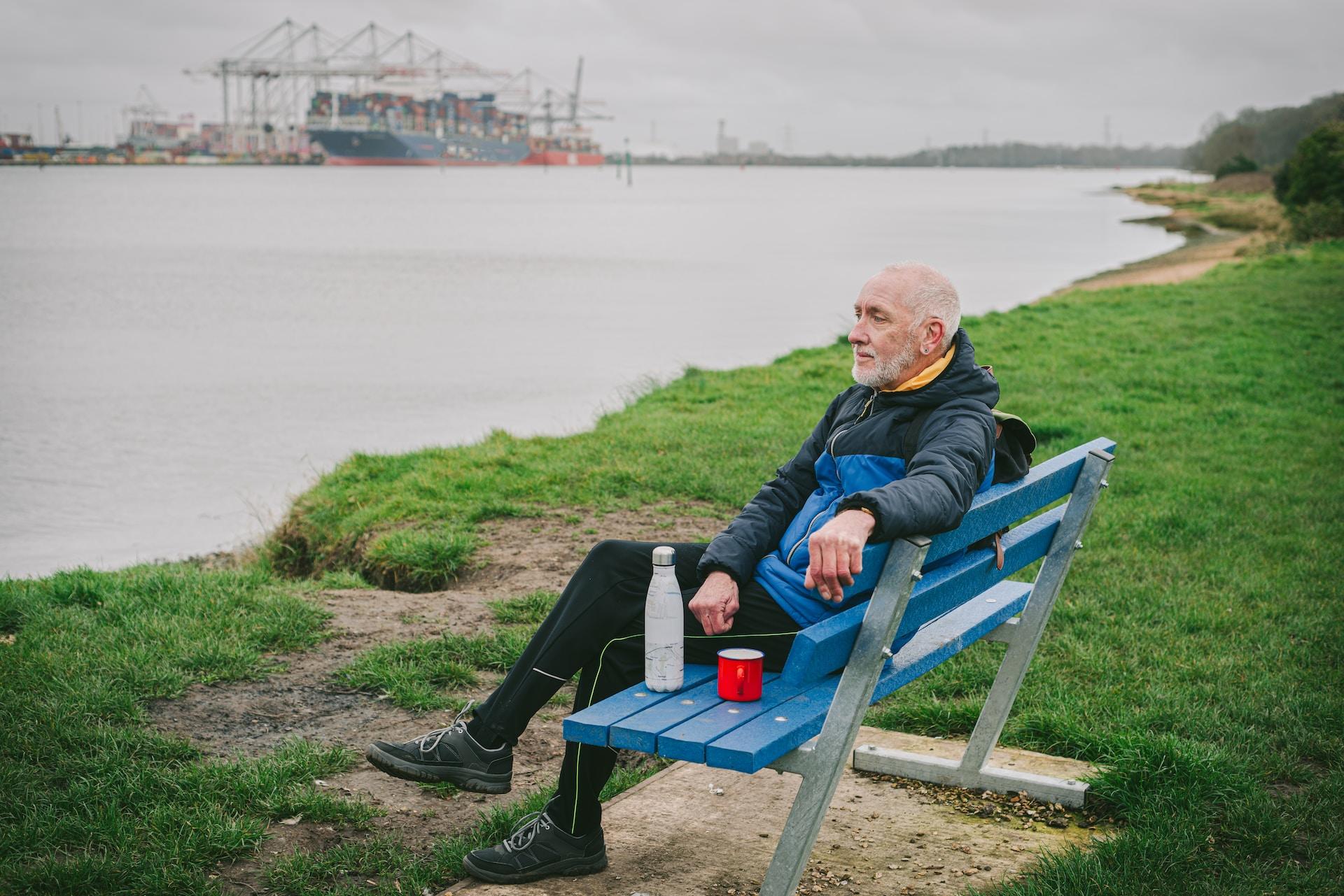 A man with short white hair and a beard, wearing a blue and black jacket and black trousers, sits on a blue bench by a body of water, with a thermos bottle and a red tin cup next to him. 