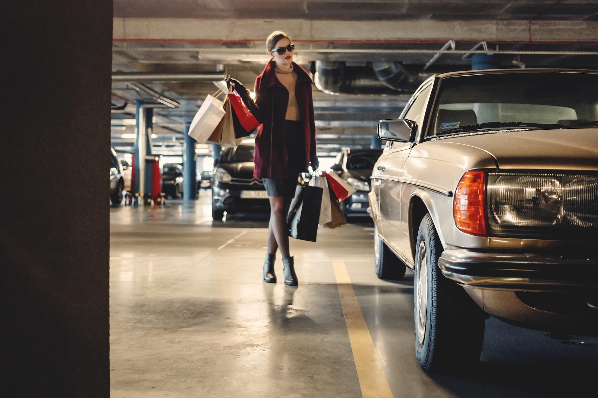 A stylish person walks towards a gold coloured Mercedes wearing a burgundy coat, a black pencil skirt and sunglasses, carrying several boutique bags in each hand. 