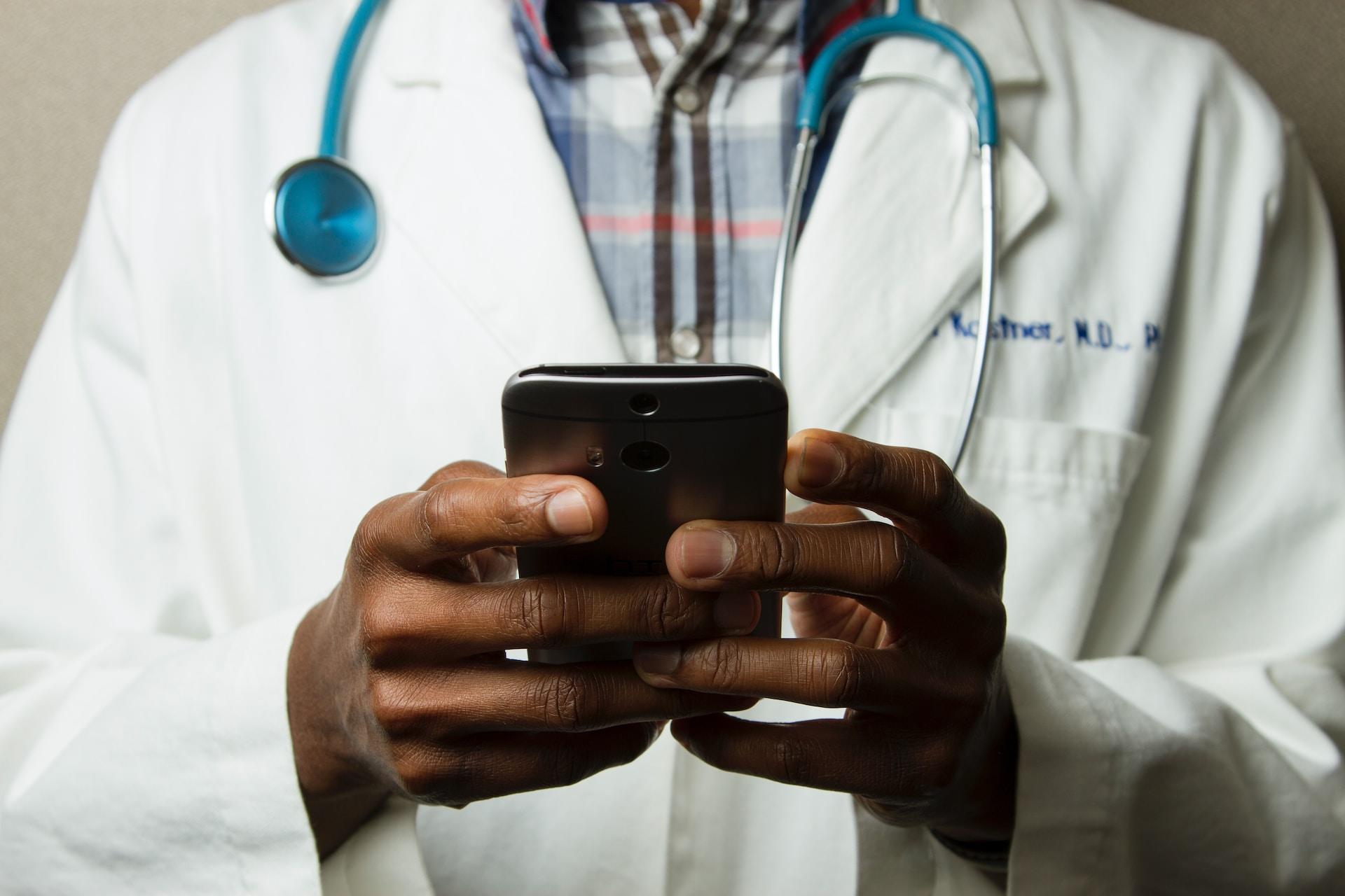 A person wearing a white lab coat over a plaid shirt, with a blue stethoscope around their neck, holds up a smartphone and works its keyboard.