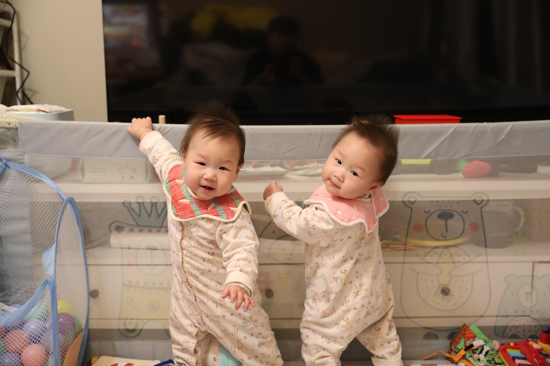 Two babies with Asian features dressed in white flowered rompers and wearing red and pink bibs stand in their playroom, holding on to a shelf.