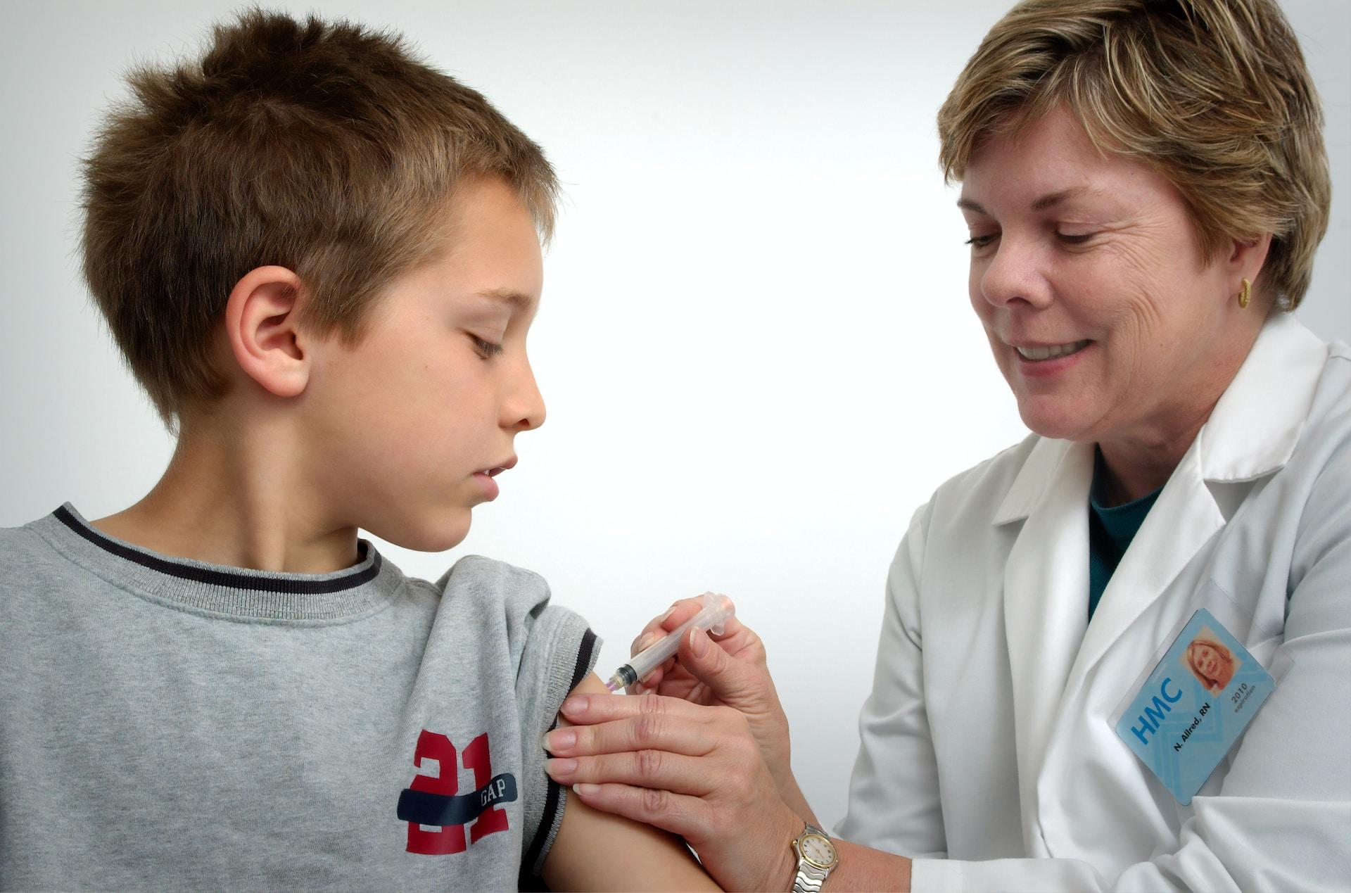 A nurse wearing a white lab coat with a green emblem on the left side smiles as they administer a vaccine to the left shoulder of a young child wearing a grey top with a dark collar, who looks on anxiously.