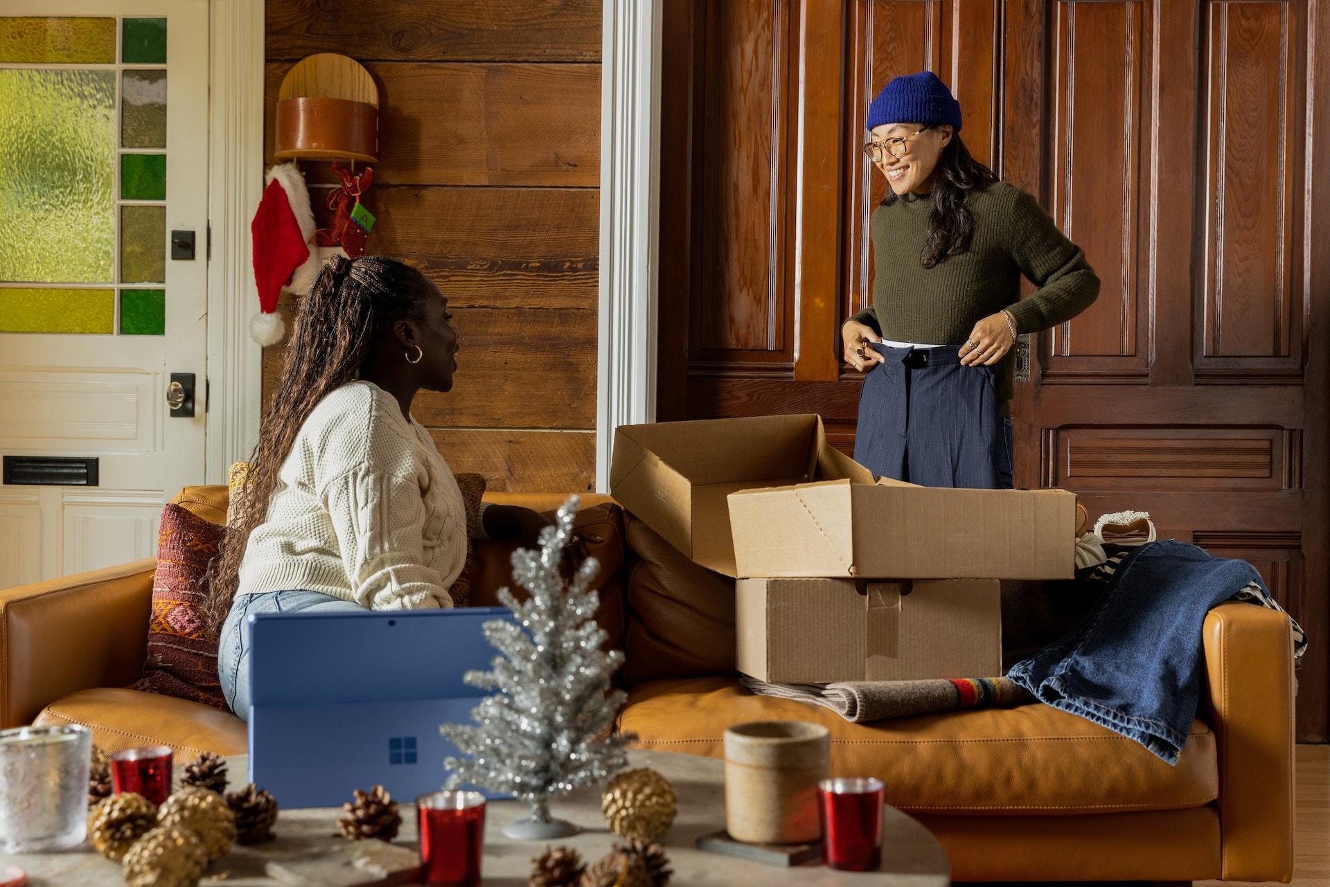 Two friends enjoying a laugh. One is standing and holding a pair of trousers in front of her while the other sits of the sofa next to open cardboard boxes.