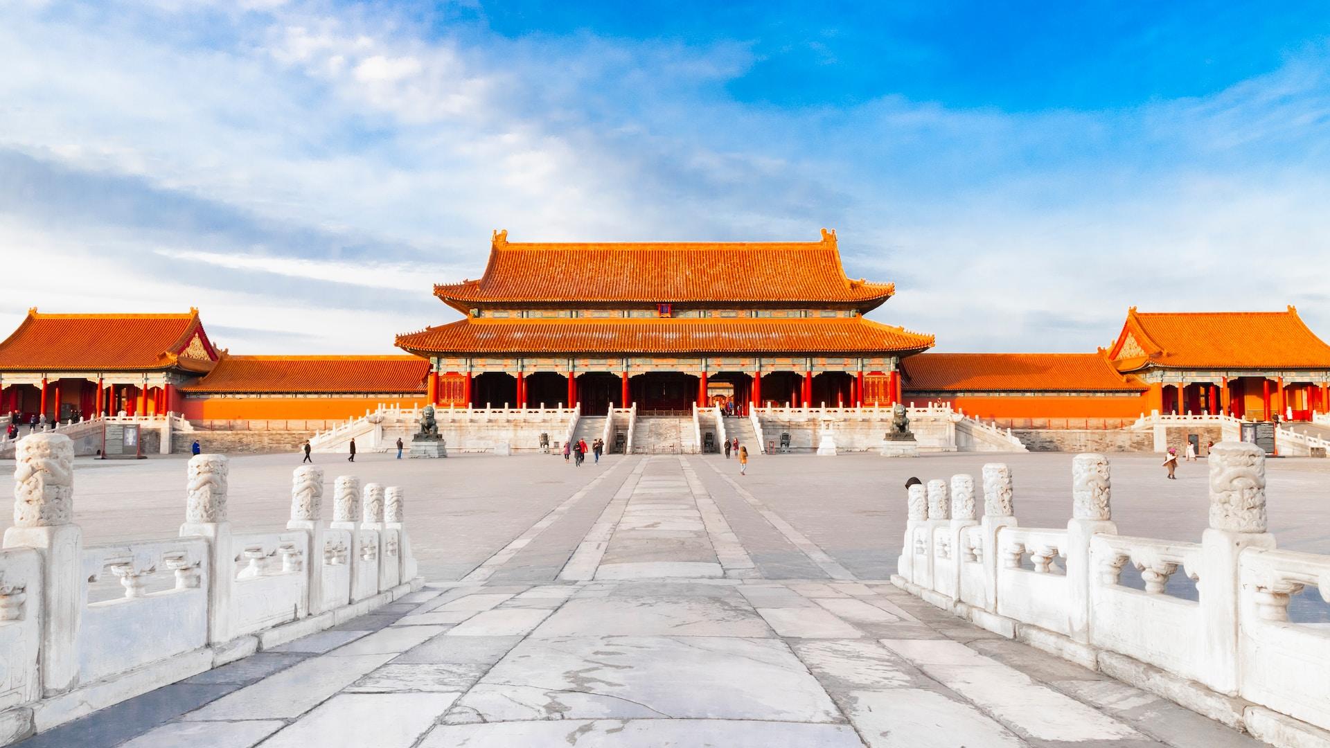 A panoramic view of the Forbidden City in Beijing on a mildly cloudy day with a blue sky visible. This courtyard has a few people ambling about. 