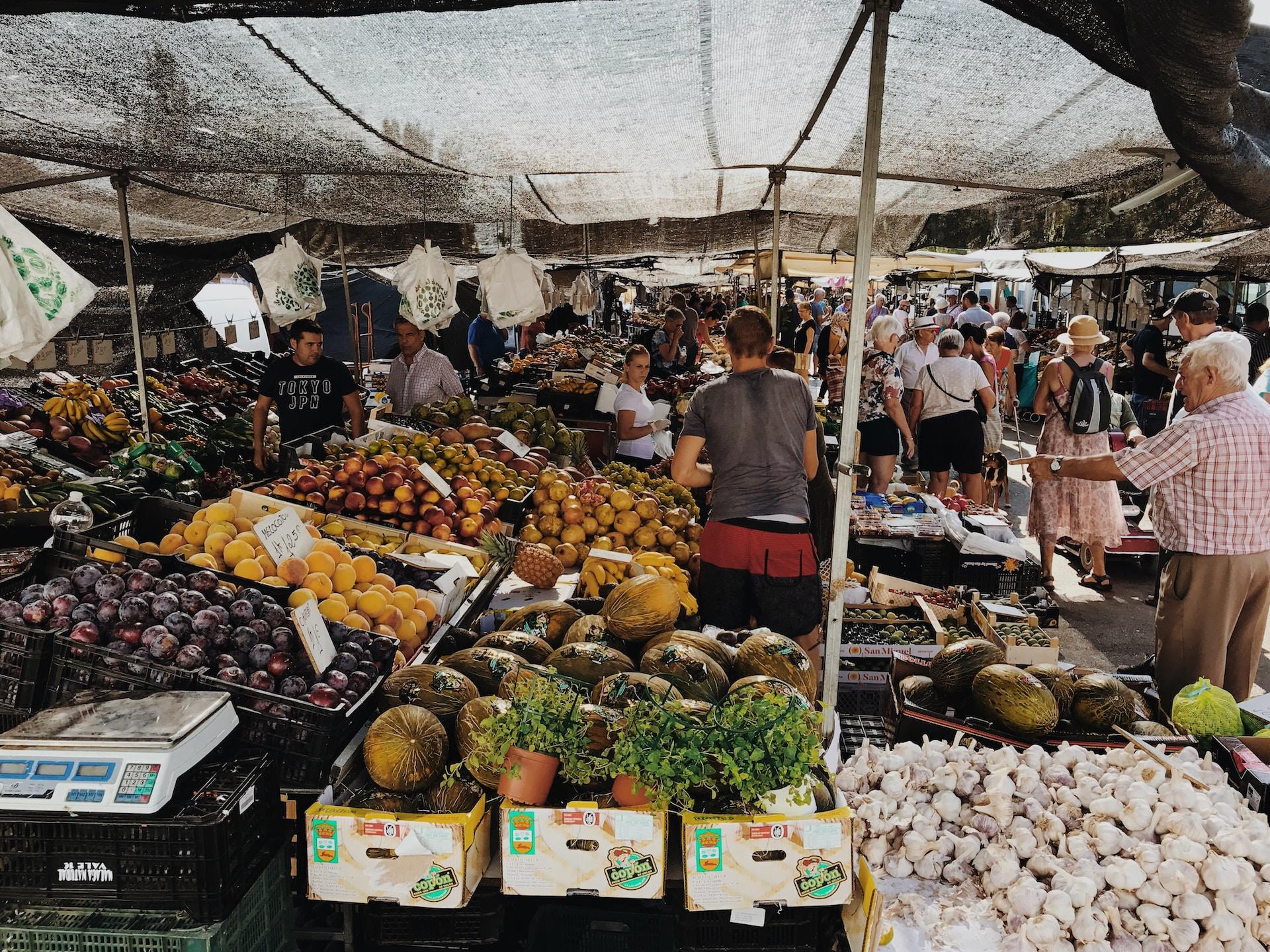 An open-air market displaying much fresh produce, with shoppers ambling down the aisles between stalls, with some stopping to buy something.
