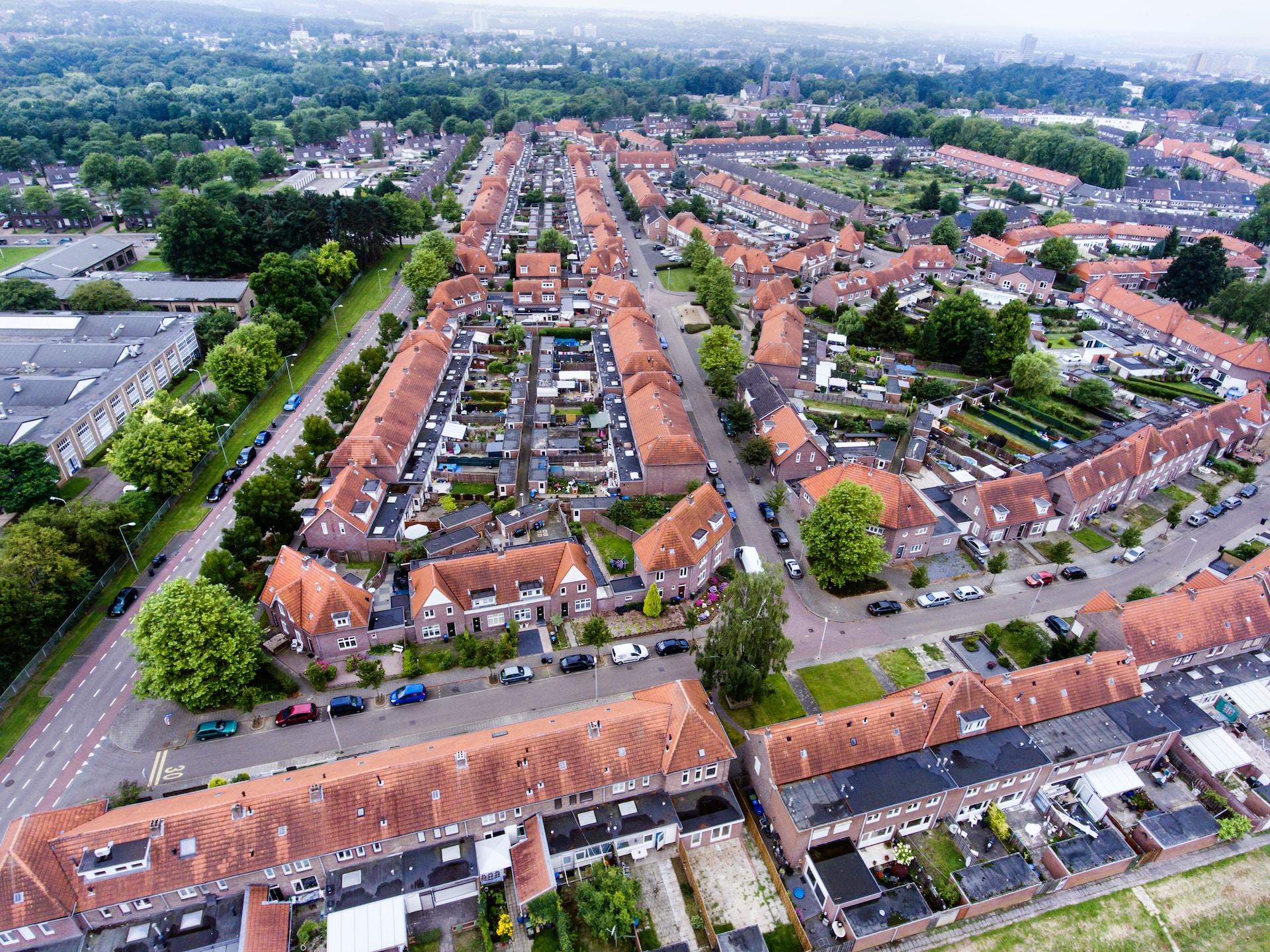 An aerial view of a housing development, featuring two-storey row houses with red tile roofs outside of an urban centre, with green trees sparse across the estate.