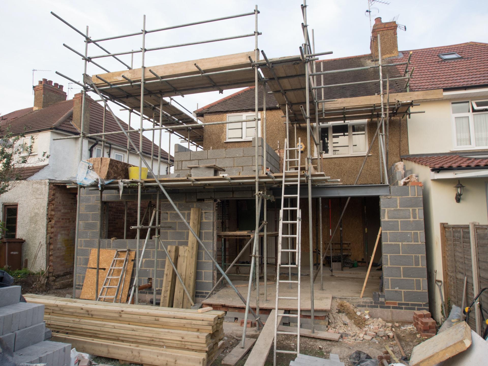 A finished home under renovation or expansion, with new grey cinderblock construction visible under scaffolding and a ladder giving access to its upper level.