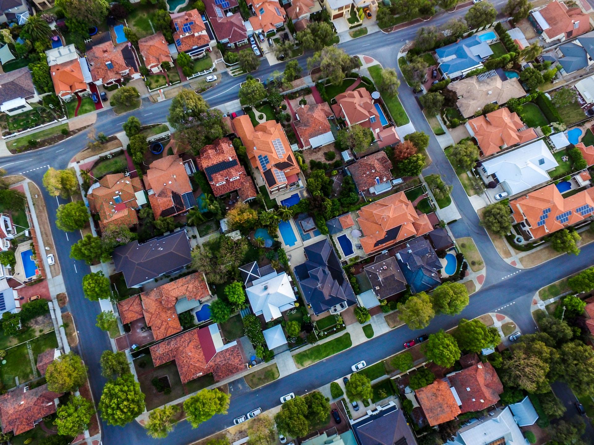 A housing estate seen from above, with many tile roofs bearing solar panels and some homes having pools. 