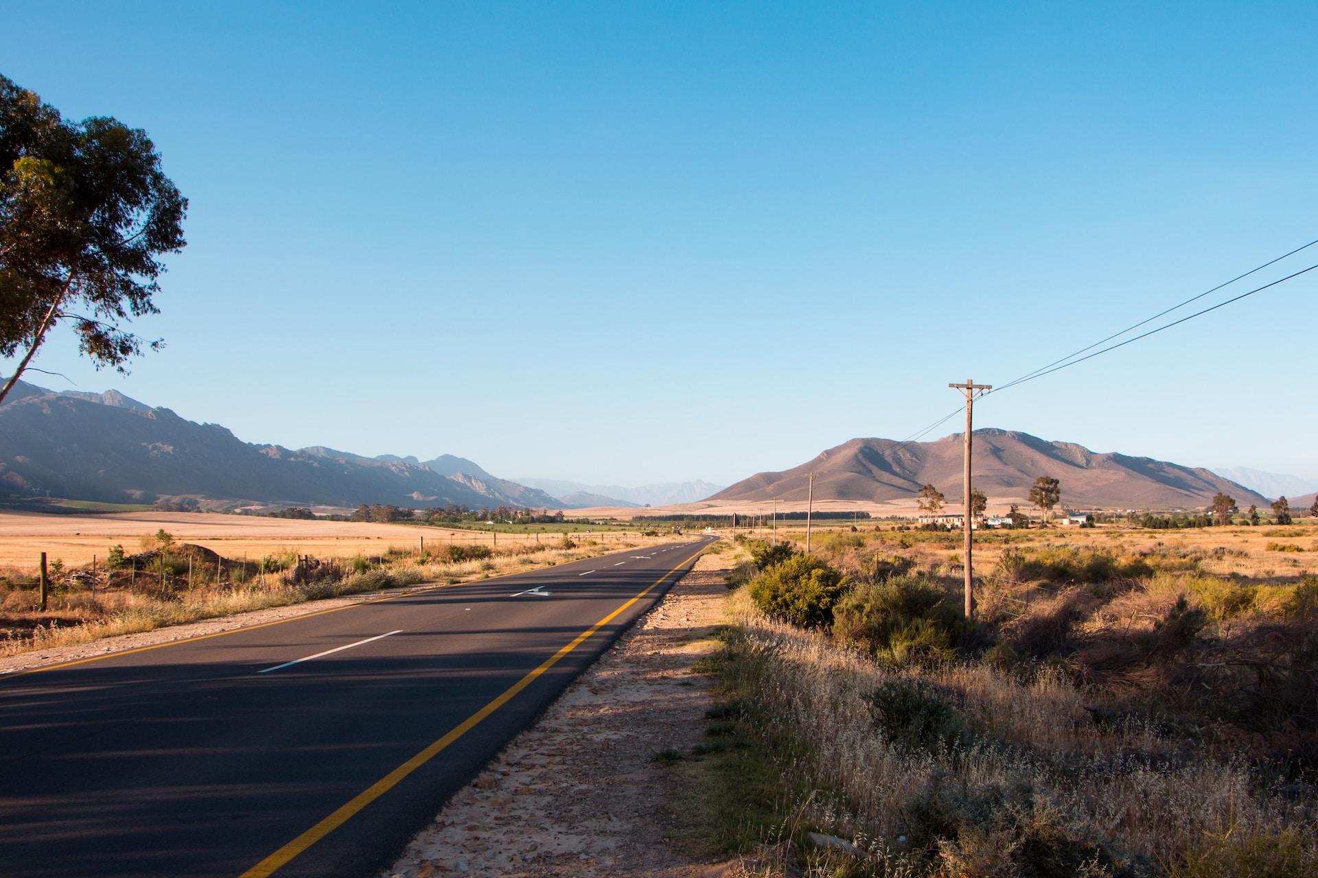 A newly-paved, deserted road lined with telephone poles leads to a distant village nestled at the foot of a mountain range. 