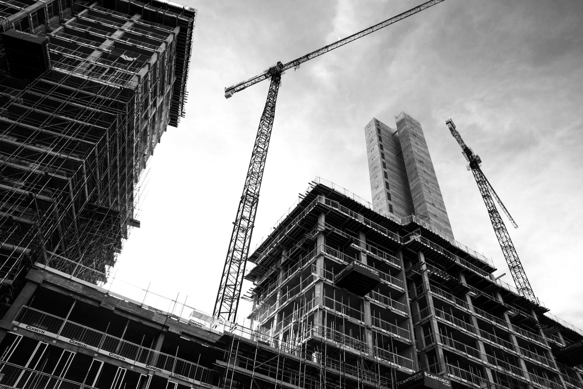 A greyscale image of a high-rise building under construction, with to building cranes towering over the constructions' skeletal frames.