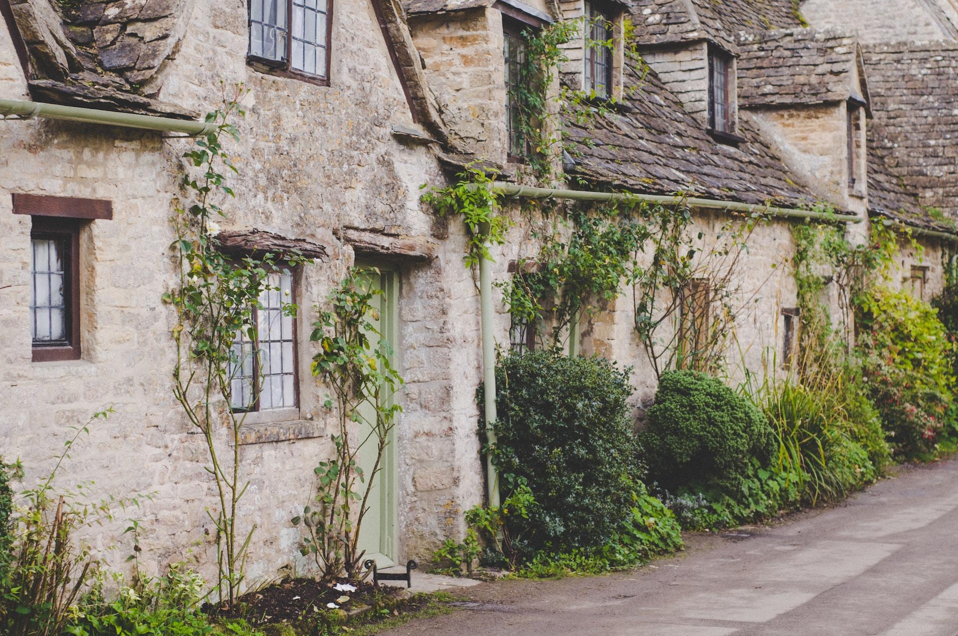 An old, dilapidated cottage build in light coloured stone with a wood shingle roof appears assailed by vegetation, with fronds climbing up the facade.