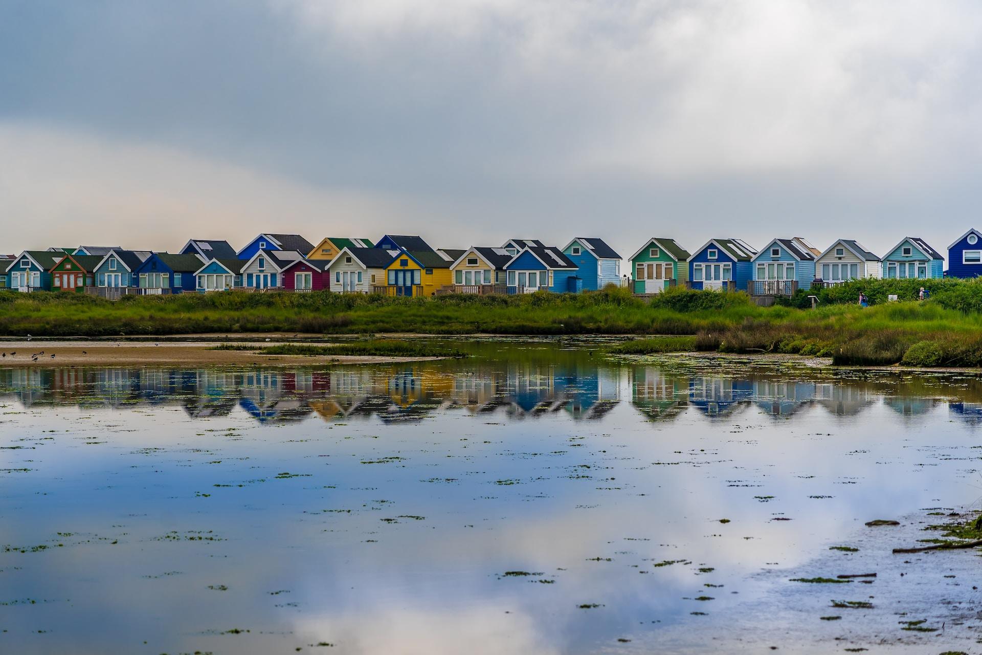A row of single-storey shanties painted in different colours facing a body of water under an overcast sky in Dorset.