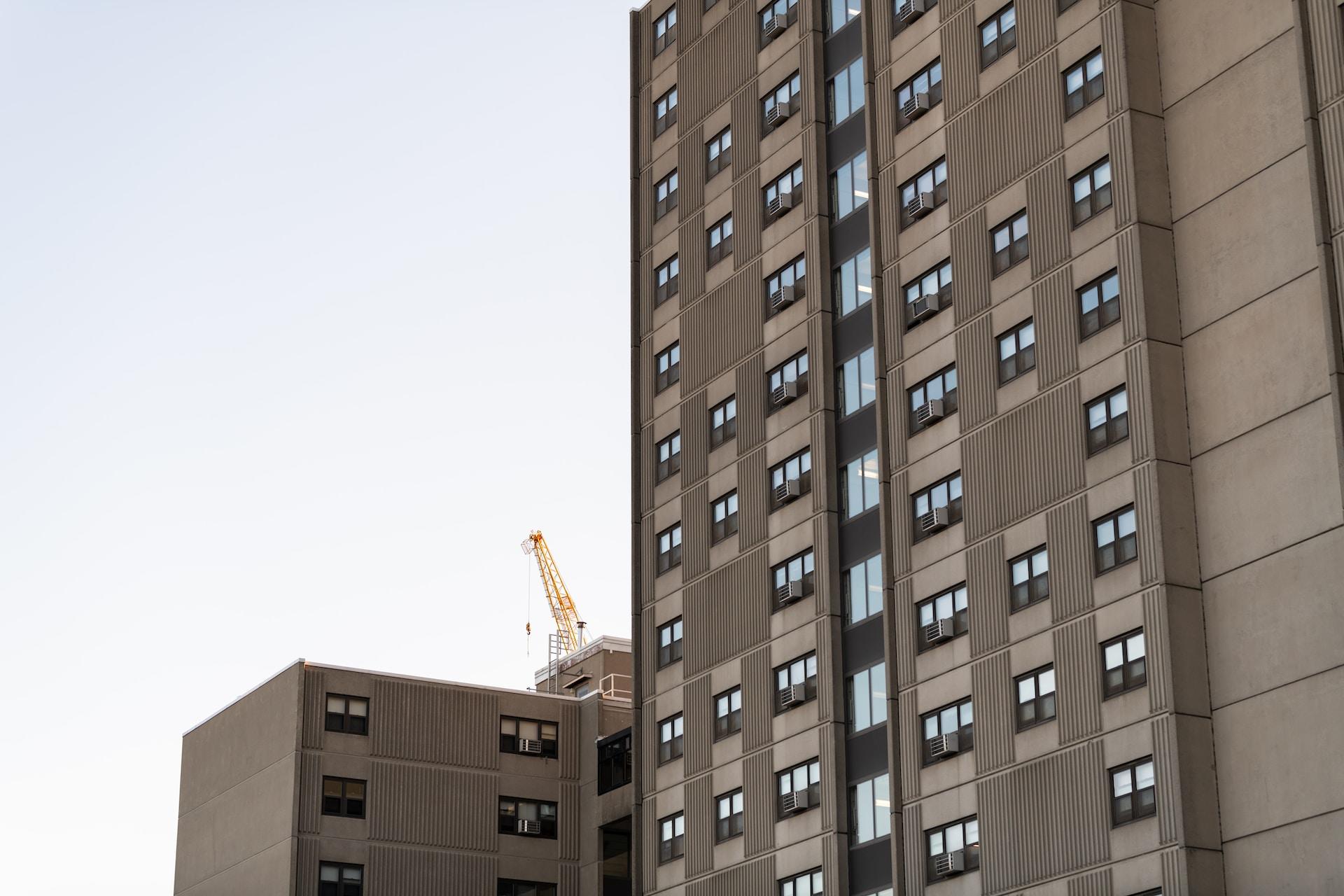 A collection of apartment blocks in dull grey tones set against a leaden sky, with a yellow construction crane visible behind them.