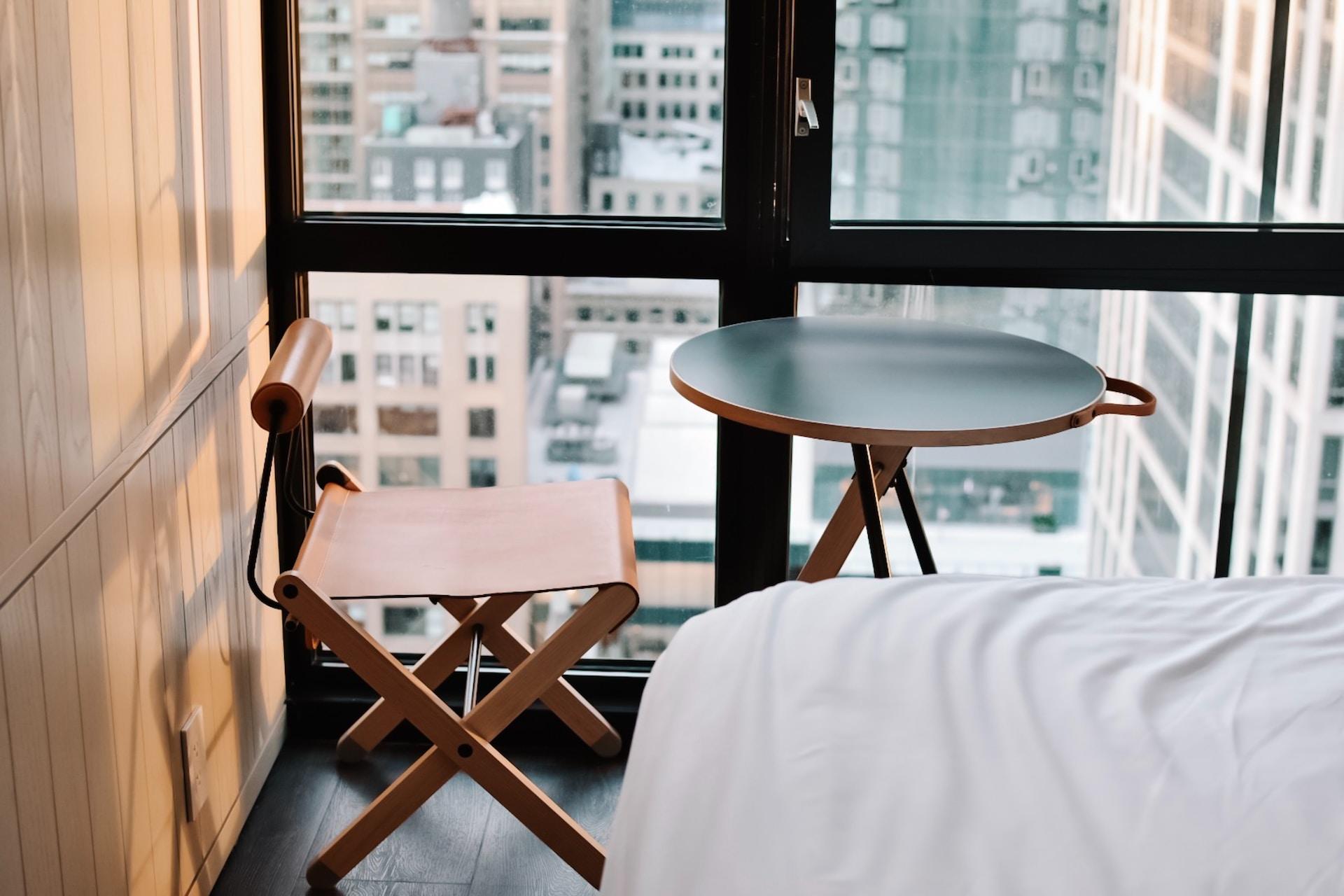 A closeup of the corner of a bed draped in white sheets, close to a designer chair made of wood and brown leather, placed in the corner, in front of a round folding table with a small green top, next to a window. 