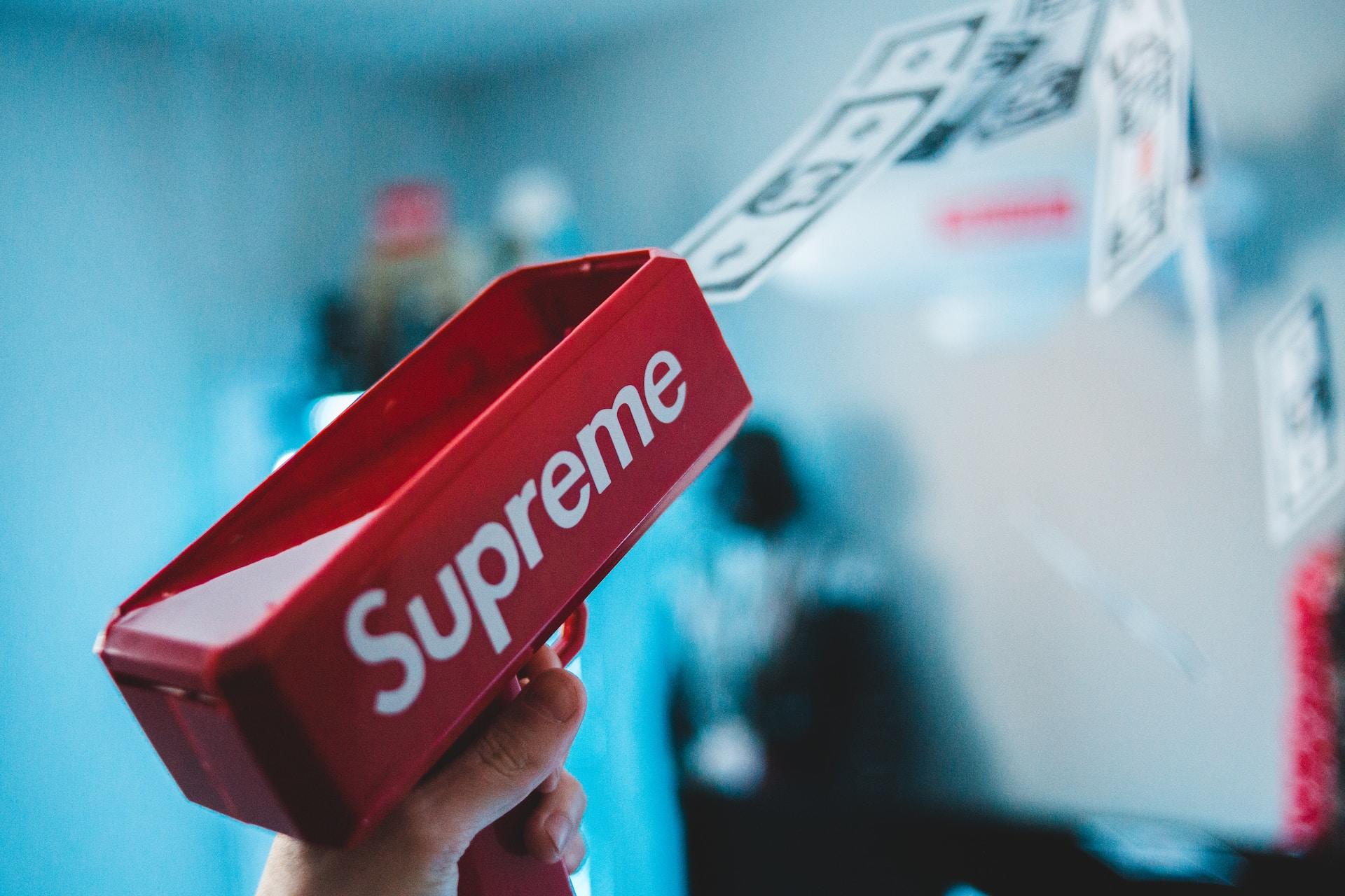 A red 'money gun' with the word Supreme written on its side in white letters shoots money out of its front side as a hand depresses the trigger.