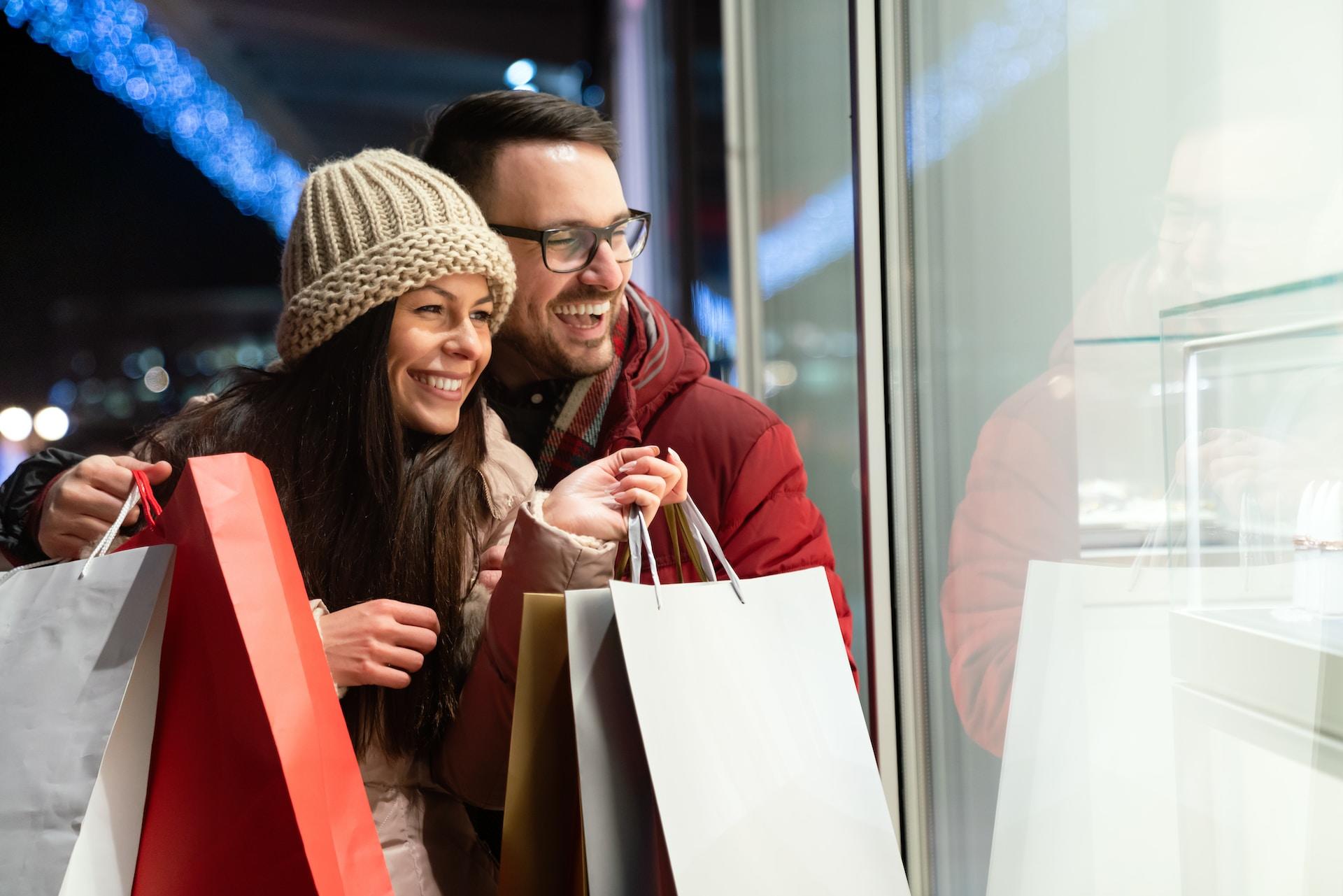 A couple wearing winter garb smile as they hold several shopping bags from fine stores and look into a shop window. 