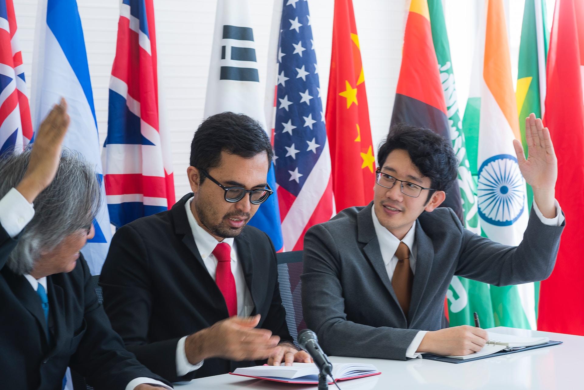 Two delegates wearing business attire raise their hands as a third delegate, seated between them, speaks while reading from an open notebook in front of them. They all sit in front of a battery of flags from different nations.