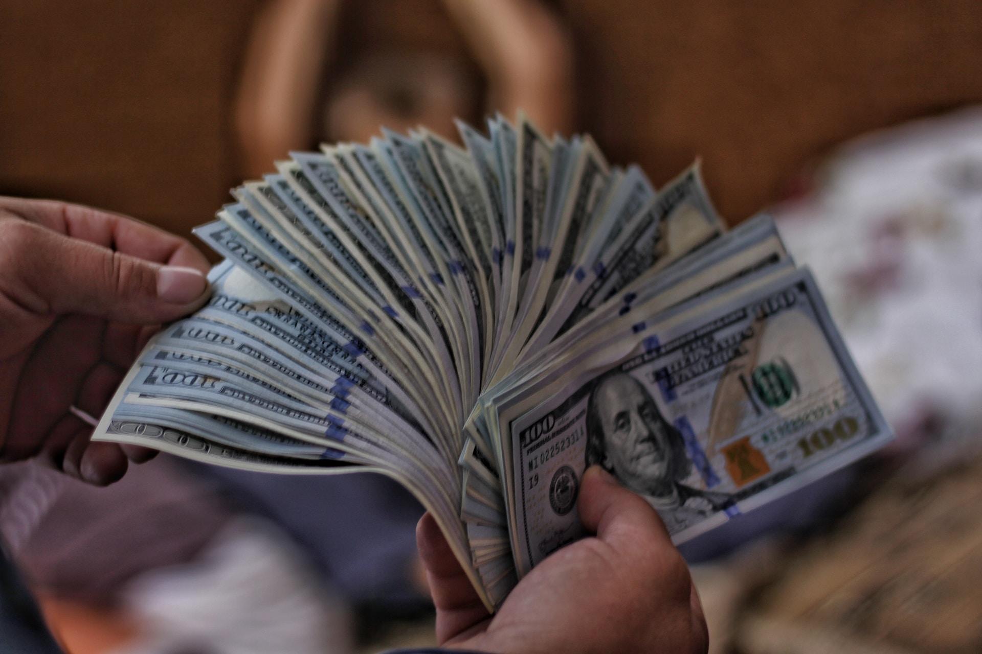 A person holding a thick wad of 100-dollar bills fanned out like playing cards in their right hand. Their left hand pinches five or six of the bills, pulling them away from the fan. 