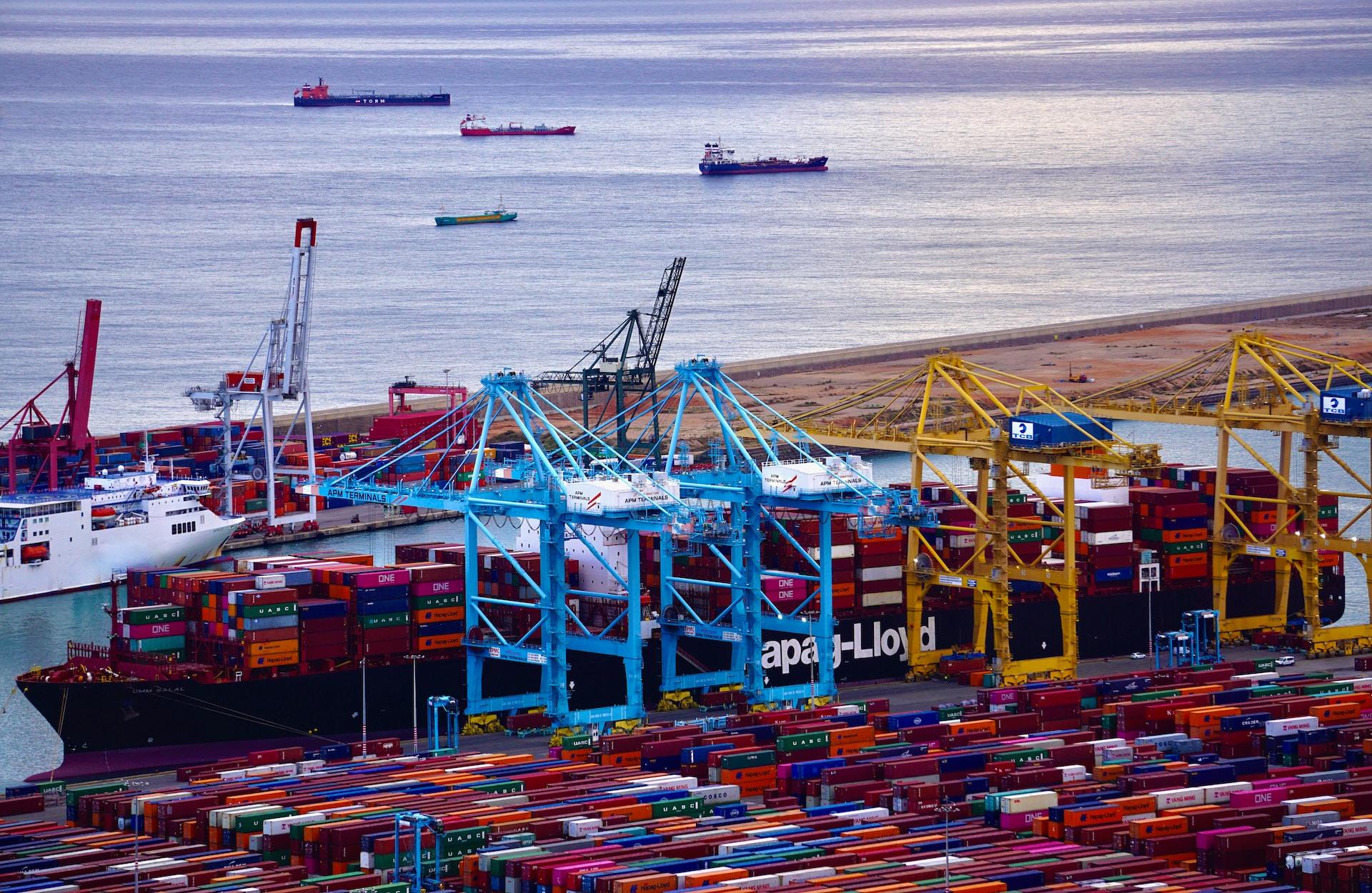 An aerial view of a shipping dock loaded with red, yellow and blue shipping containers and a body of water behind it.