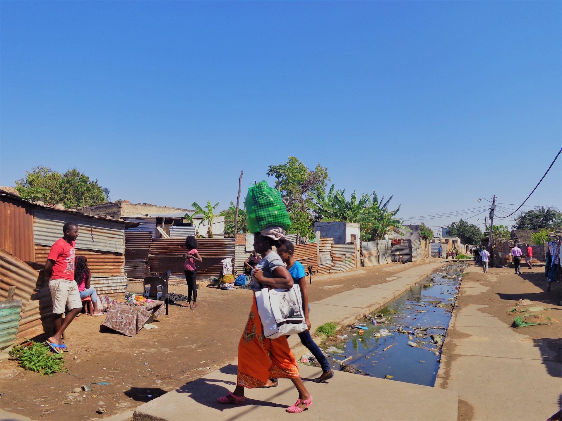 A view of Maputo, Mozambique, with a concrete slab spanning a narrow canal with debris floating in it and two people walking across the slab. The image shows corrugated tin shacks lining the paths on either side of the canal.