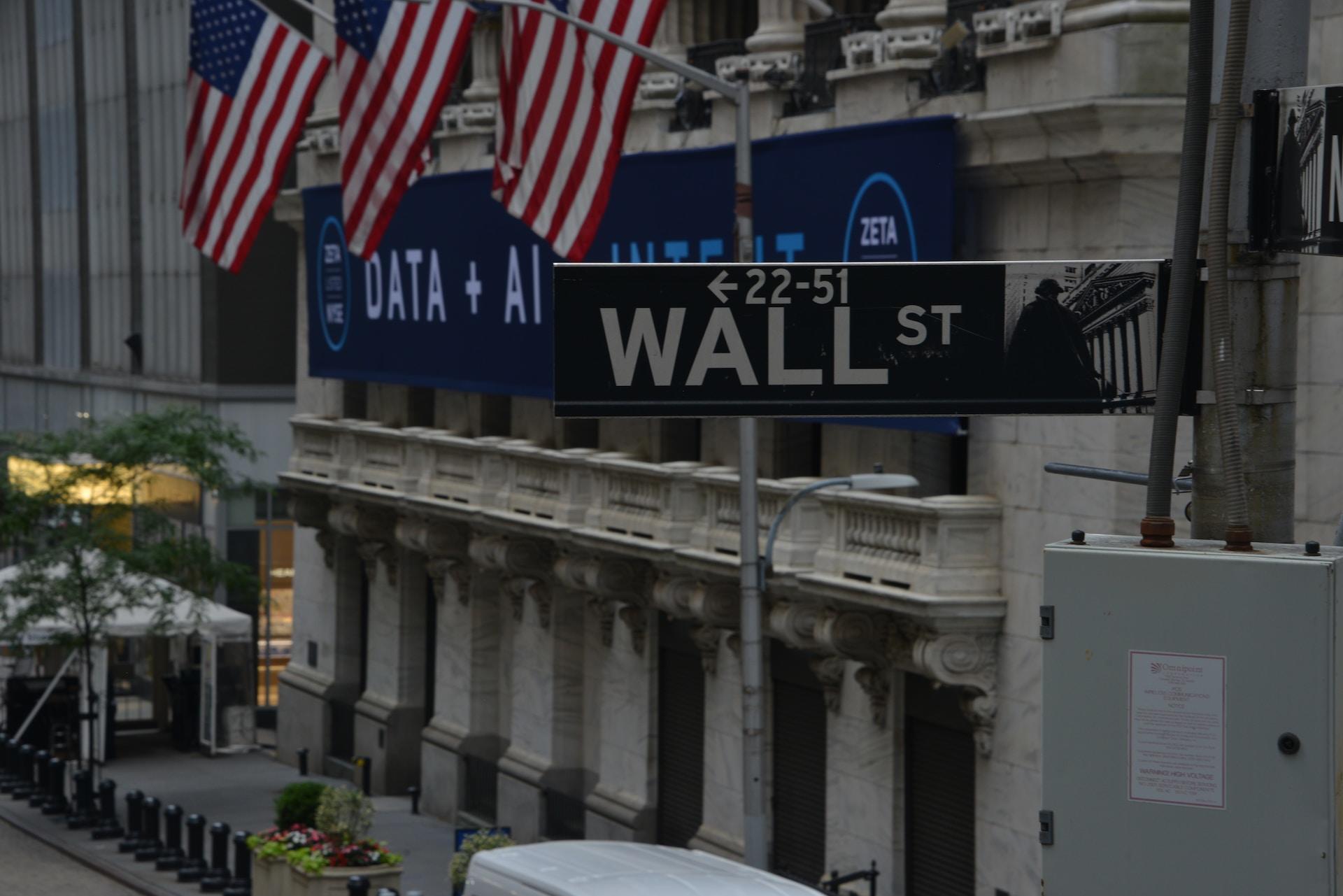 A black street sign with white lettering proclaiming Wall Street in front of a building bedecked with American flags that bears a blue sign with white lettering.