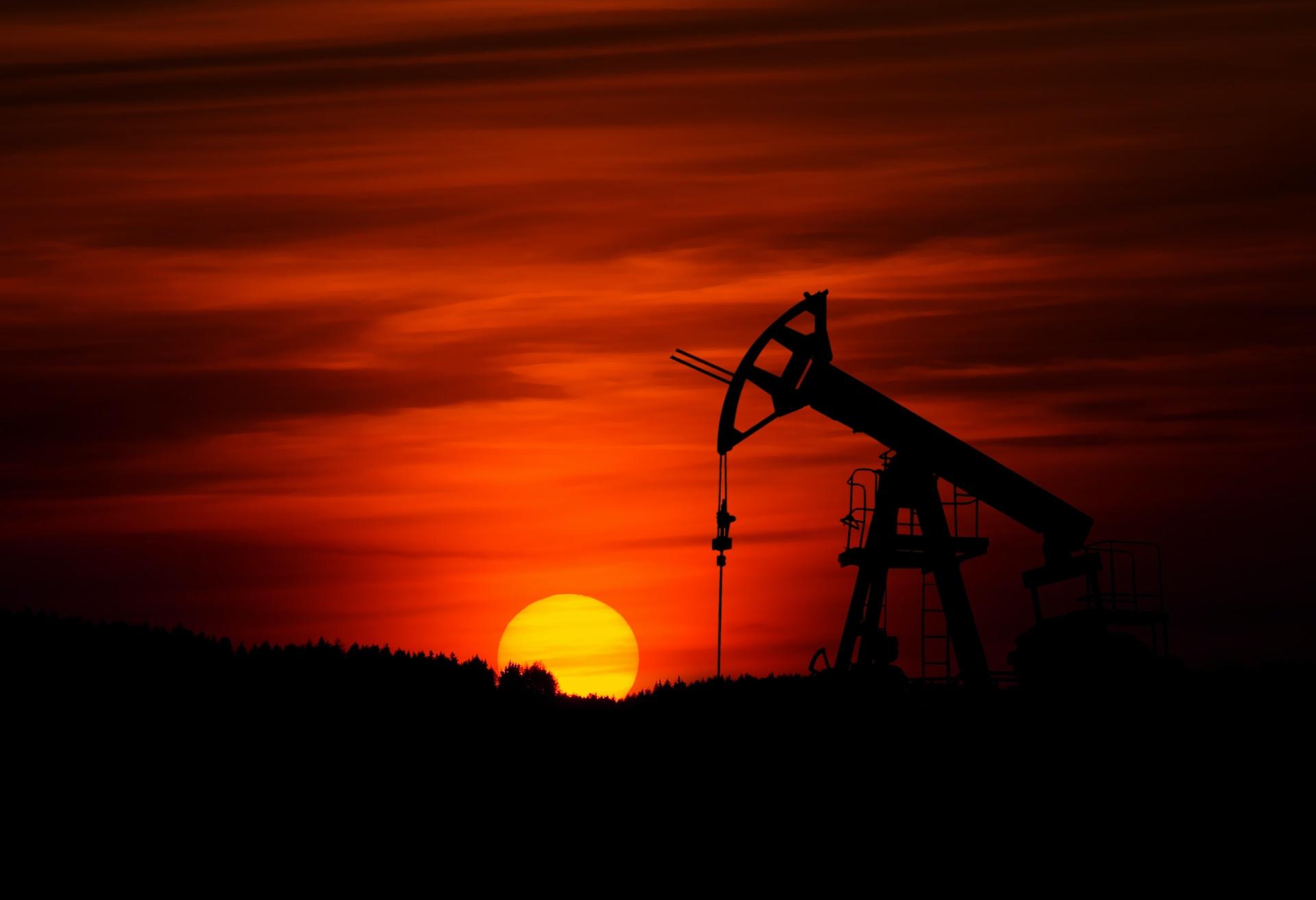 An oil derrick with its drill head in the up position, seen in silhouette at sunset, as the sky turns burnt sienna colour and the landscape is in shadow. 