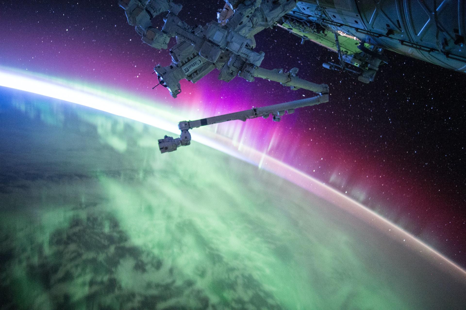 A space vehicle with a robotic arm hovers over a planet emitting a greenish light that contrasts with a mauve aurora.