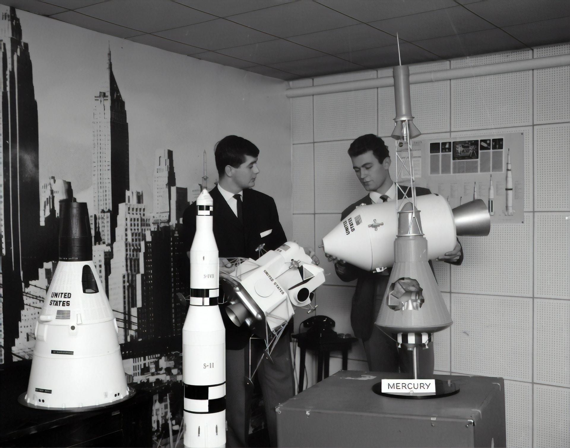 A black and white picture of American rocket and space capsule models on display, with two men in suits and ties admiring them. 
