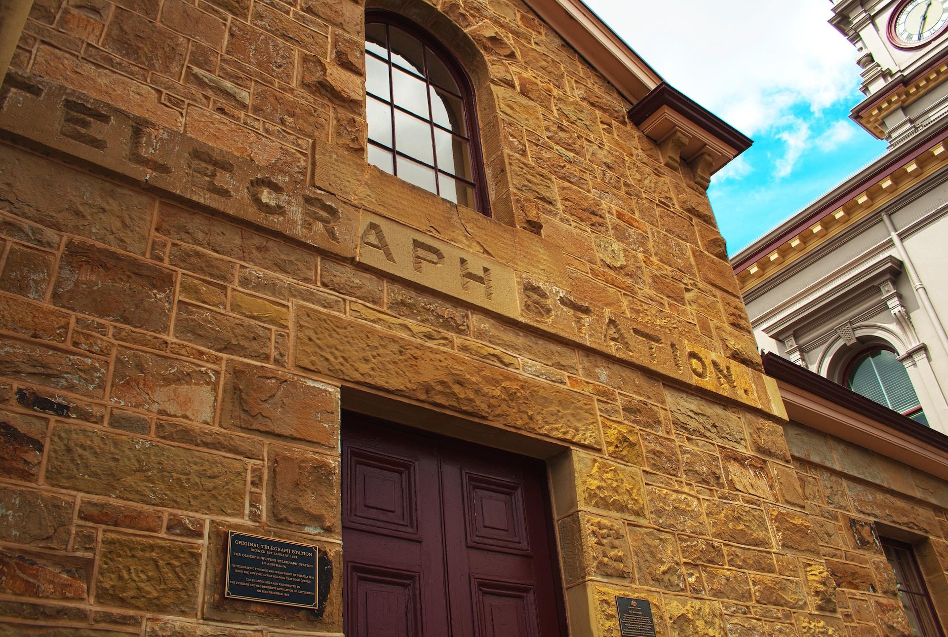 An old stone telegraph centre in Victoria Australia with brown doors and a window above it, on the second level. Next to the door is a black and brass historical facts plaque. 