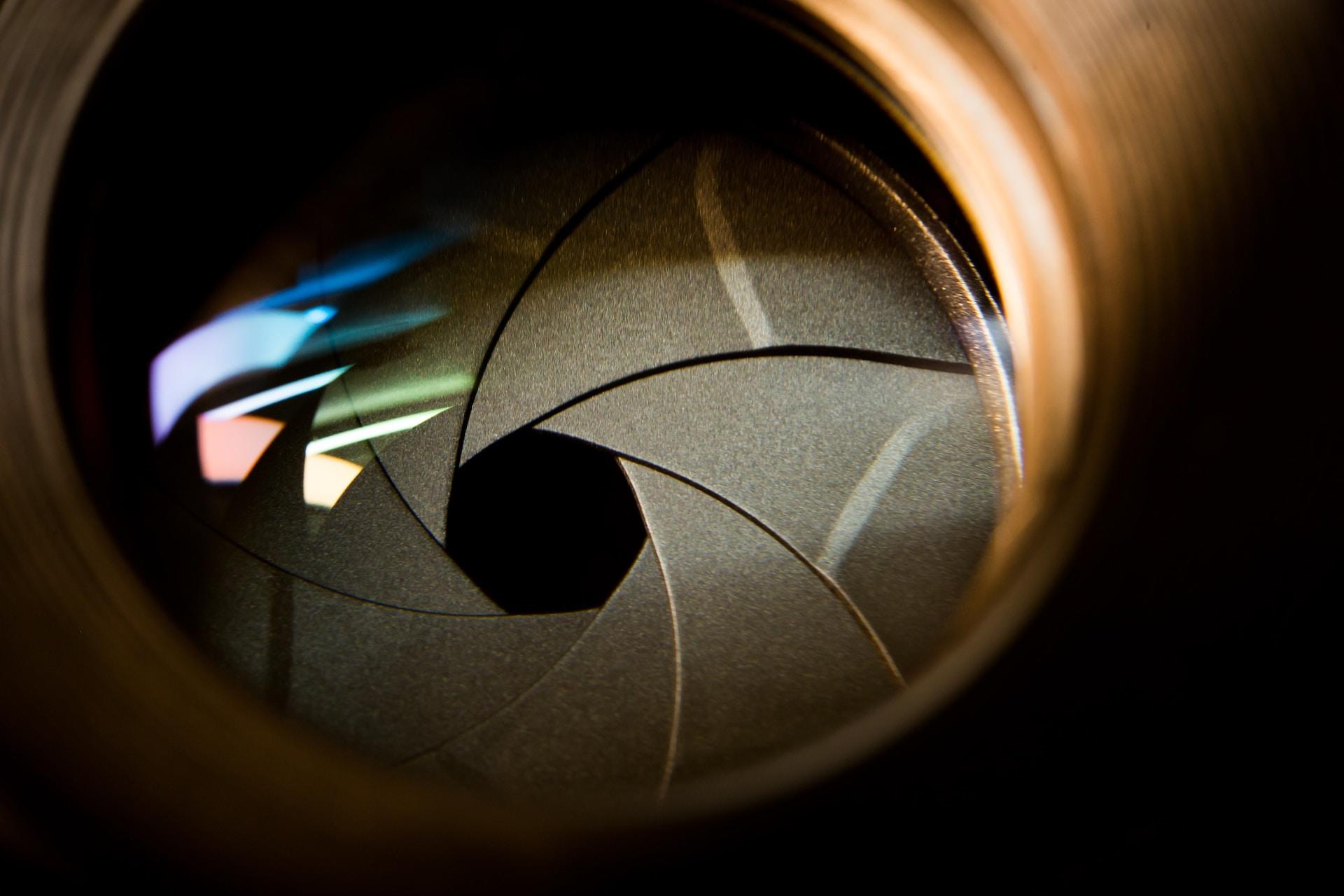 An extreme closeup of a camera's shutter aperture with three reflected bands of light on the lens; one blue, one pink and one yellow in tone.