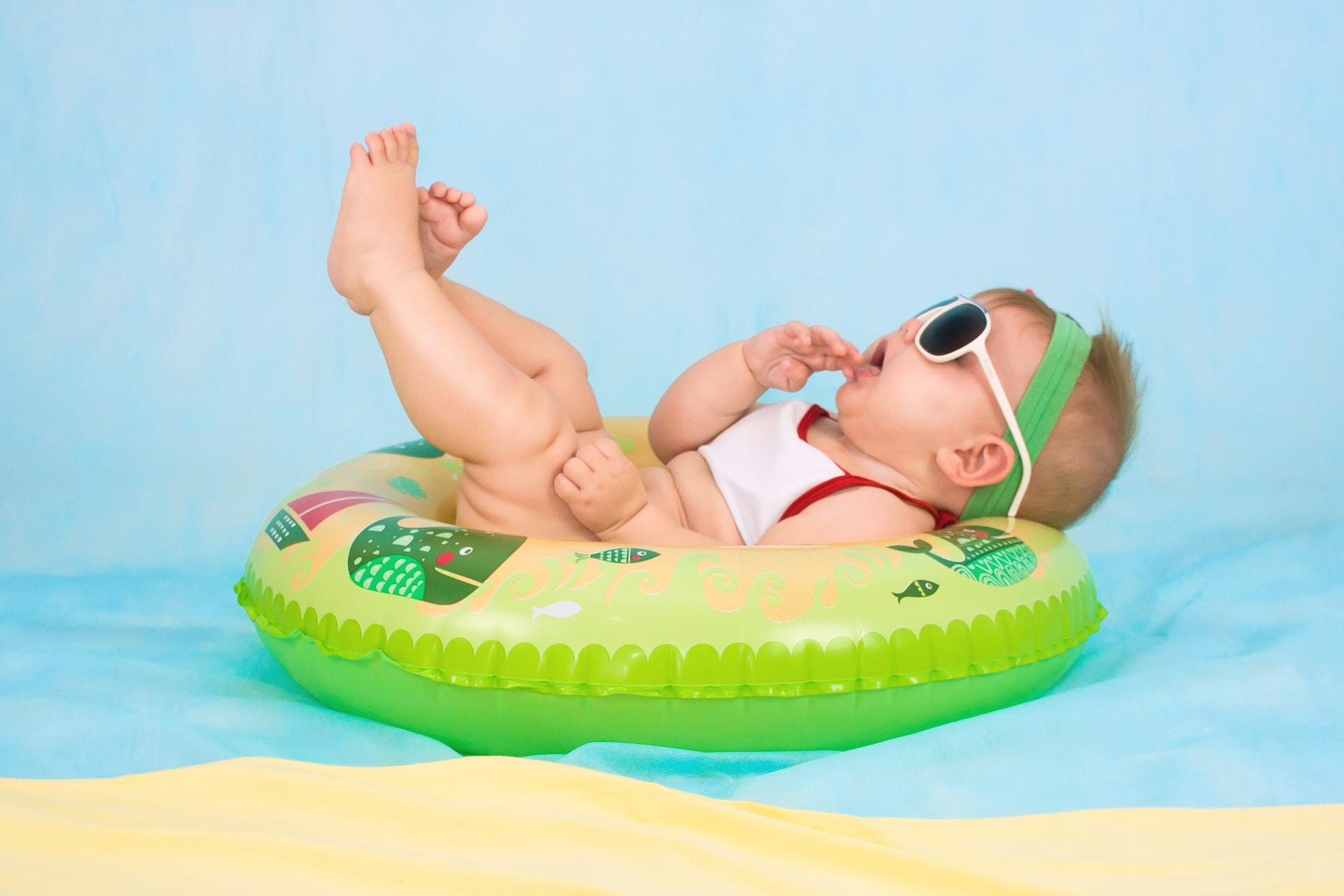 A baby wears sunglasses and a white outfit as they lay in a green pool float with their feet in the air.