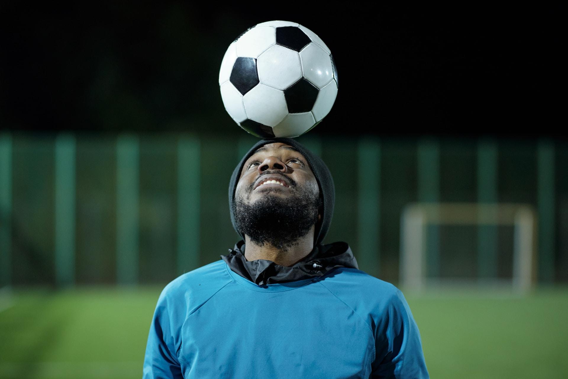 A person wearing a two-tone blue top stands with their head back, balancing a soccer ball on their forehead. 