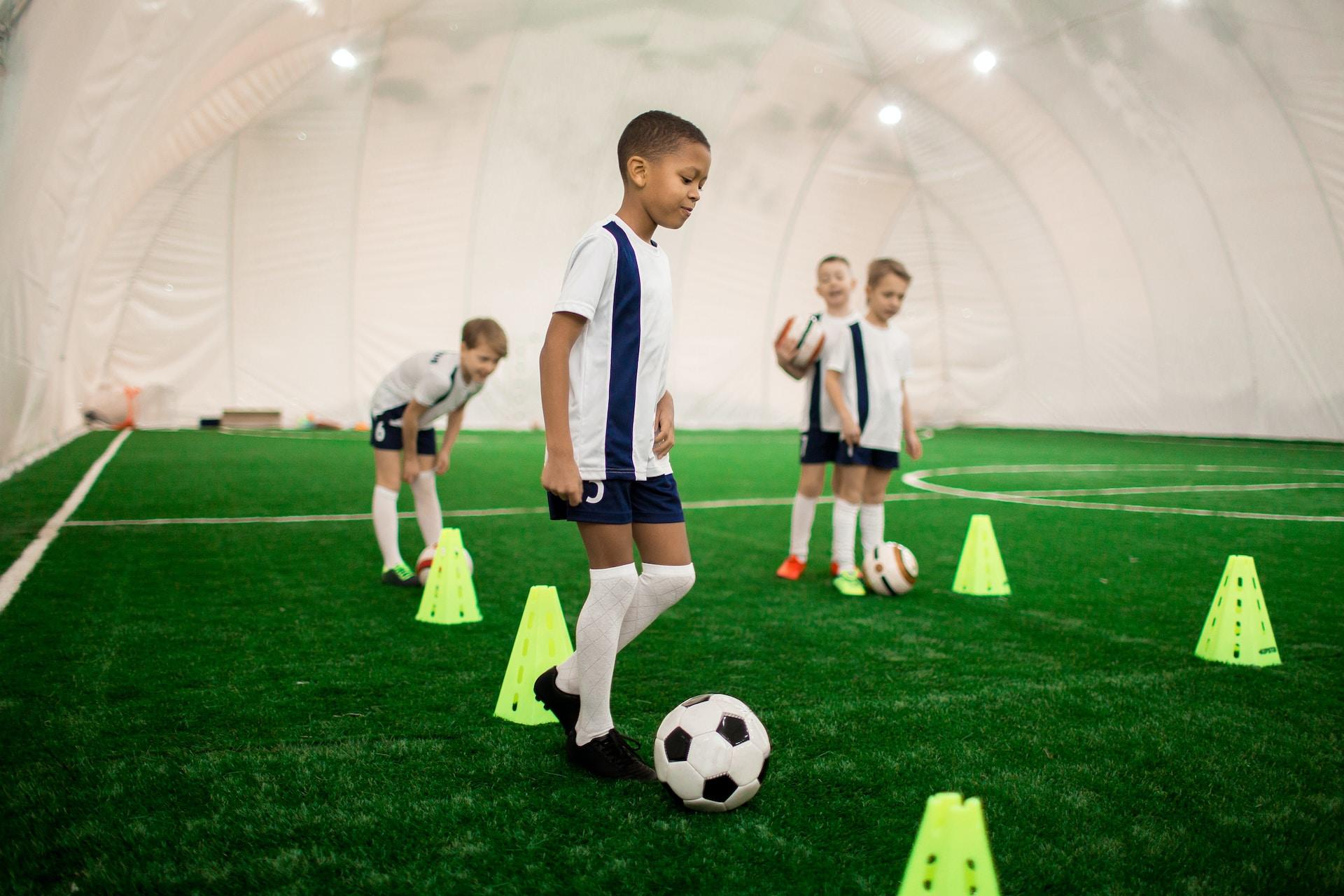 Young soccer players wearing white and dark blue uniforms stand on a field that has neon yellow cones set up on it, preparing to practise ball skills.