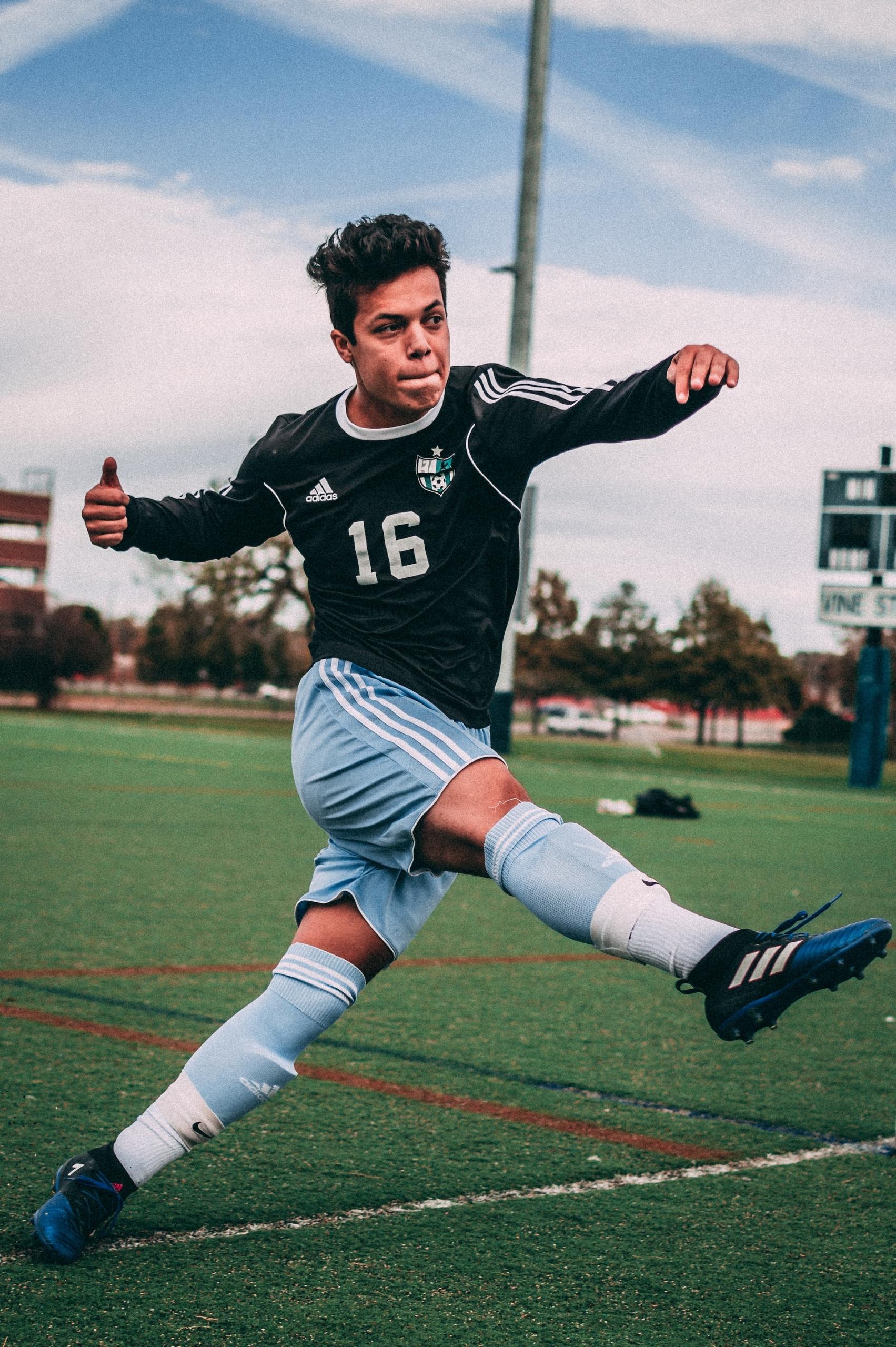 A soccer player wearing a black long-sleeved jersey and light blue shorts twists their body to deliver a powerful kick with their right foot.