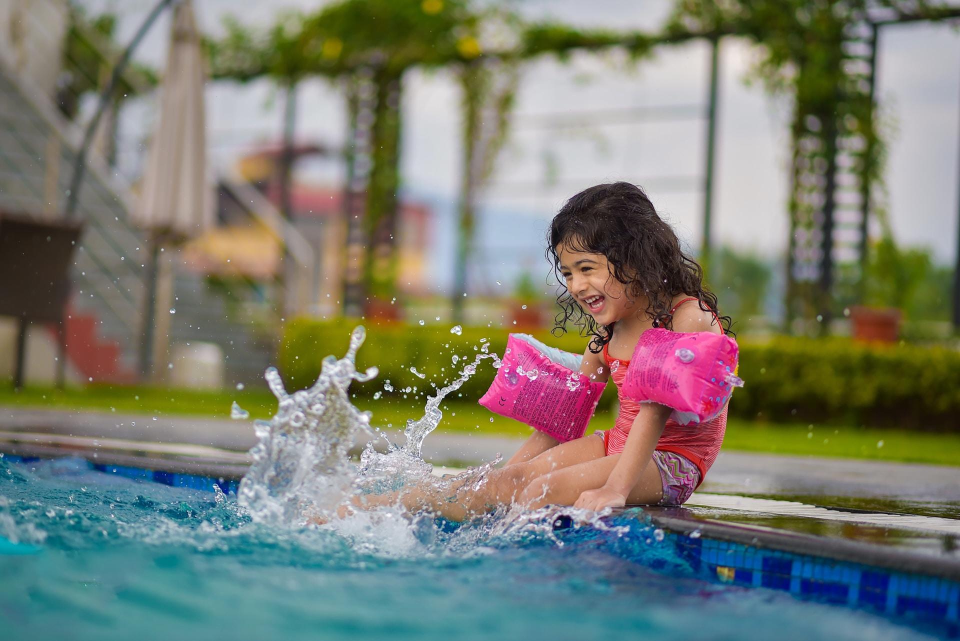 A small girl wearing a bright pink suit and matching water wings sits at the edge of the pool, kicking her legs and splashing water. 