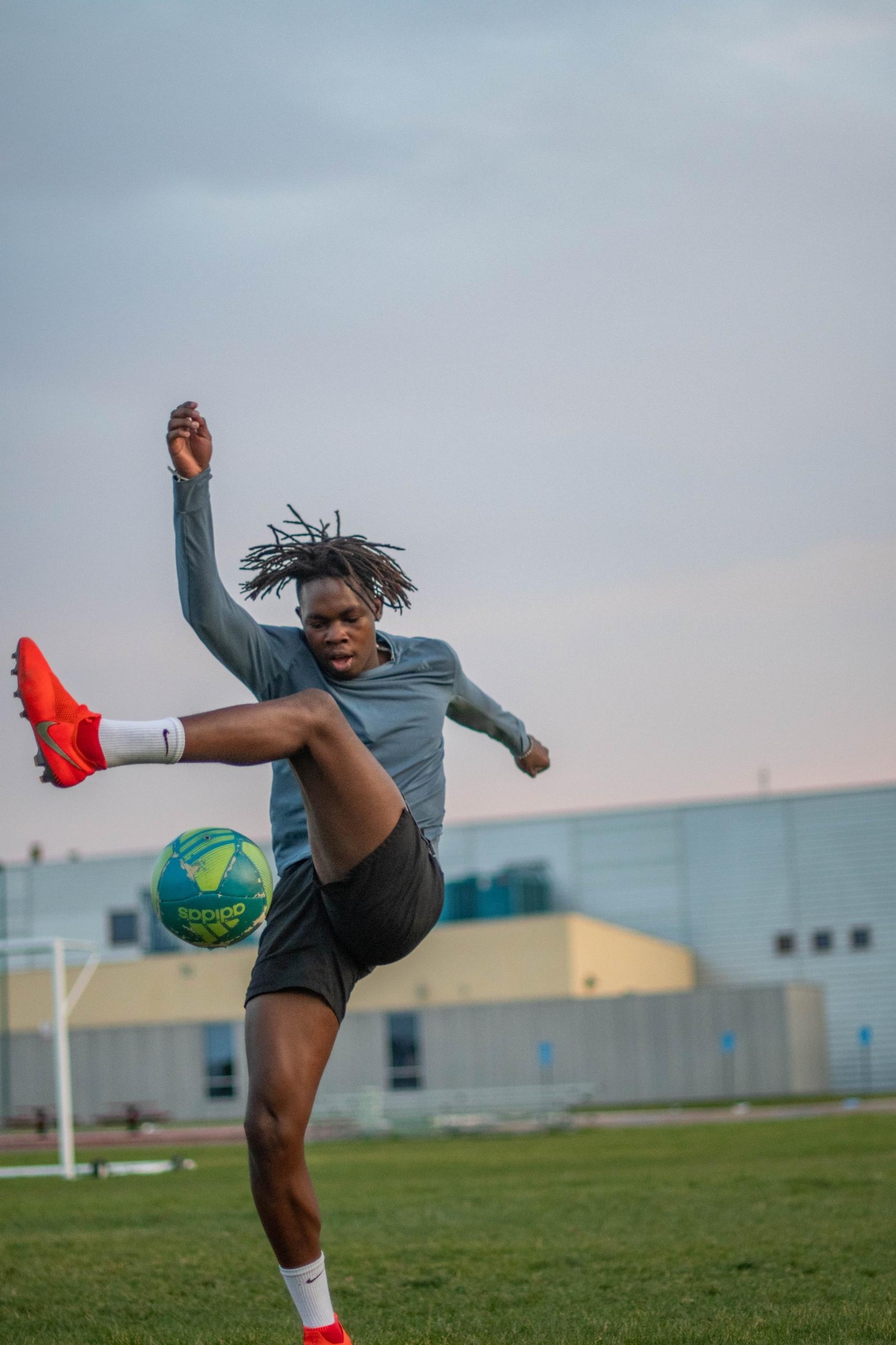 A person wearing a grey-green long-sleeved shirt, black shorts and neon orange cleats misses kicking a green and yellow soccer ball.