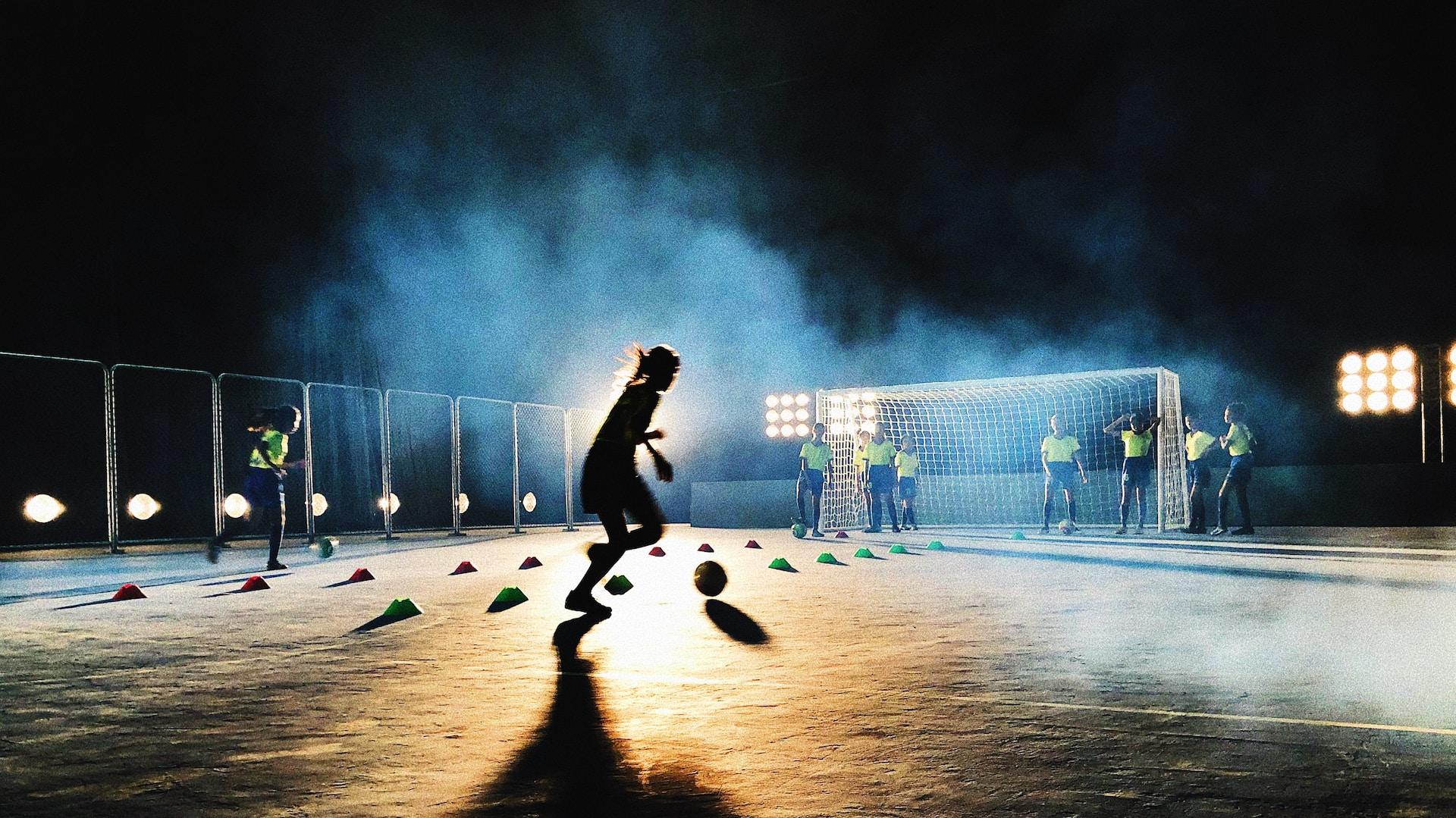 A soccer trainee silhouetted under stadium lights lines up for the kick into the goal while people watch in the background.