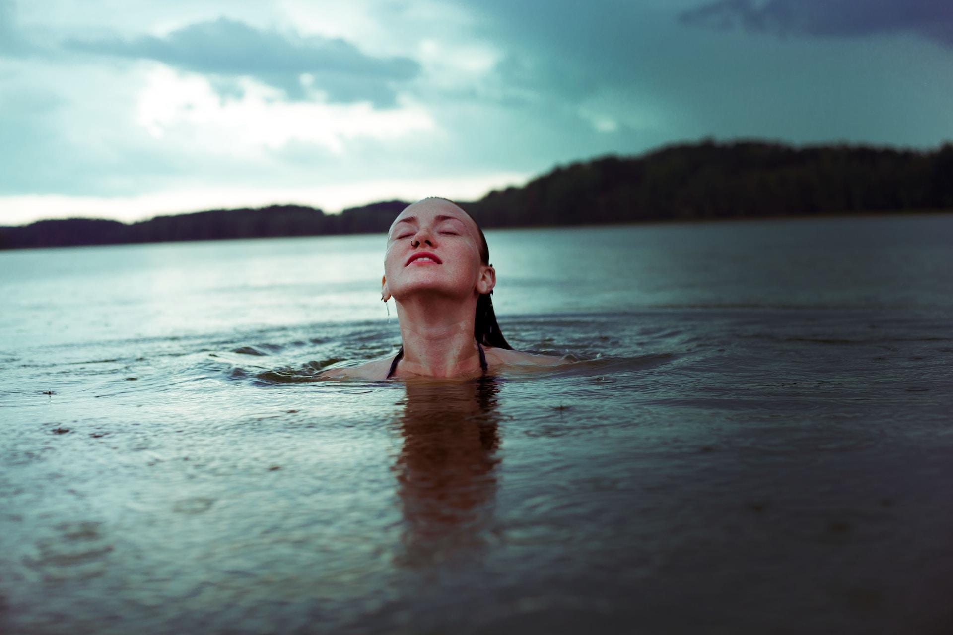 a person stands in a body of water visible only from the neck up, with a landmass visible behind them and a grey sky above. 