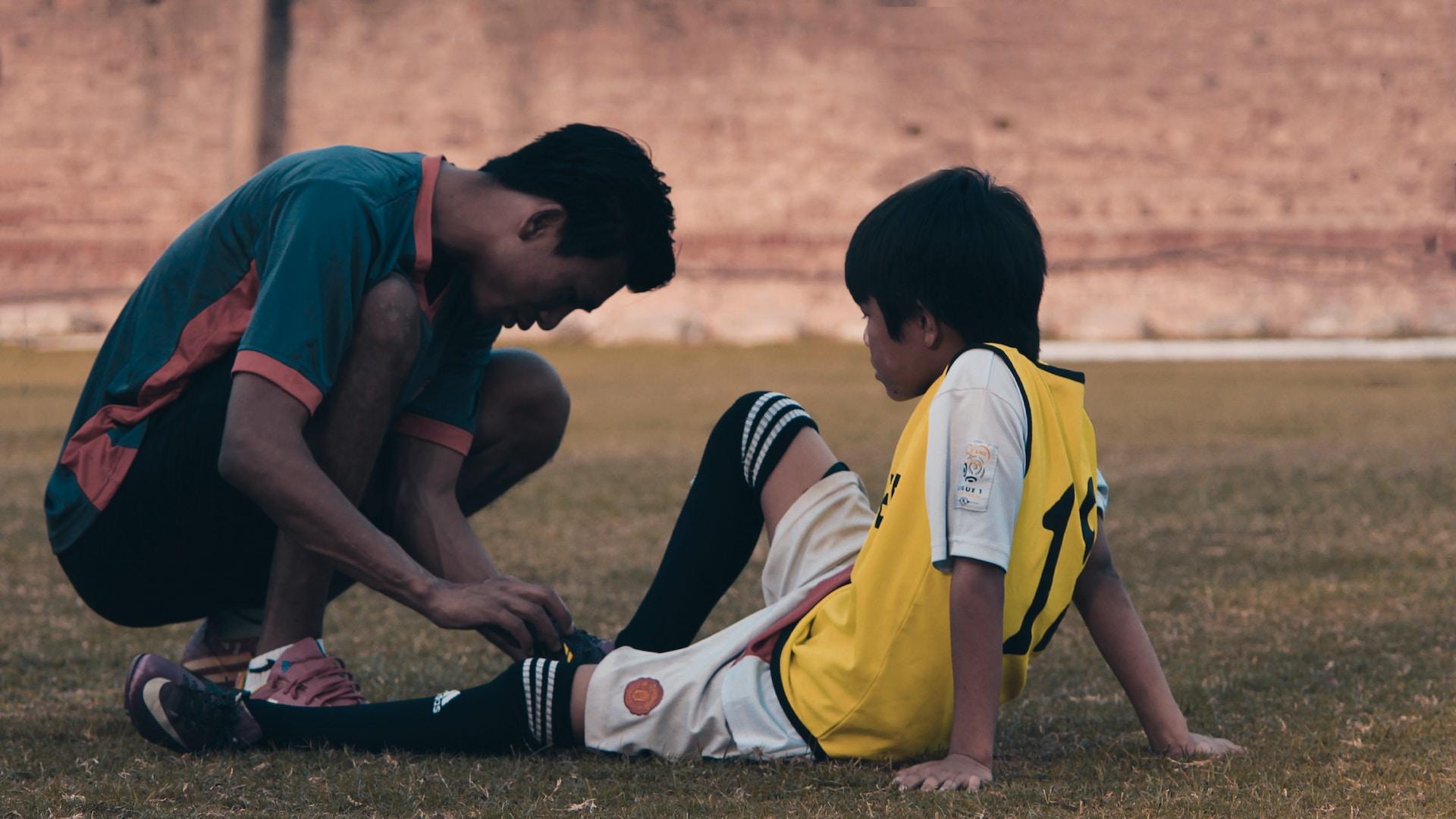 An adult kneels in front of a child wearing yellow and white soccer gear, tending the to child's knee.