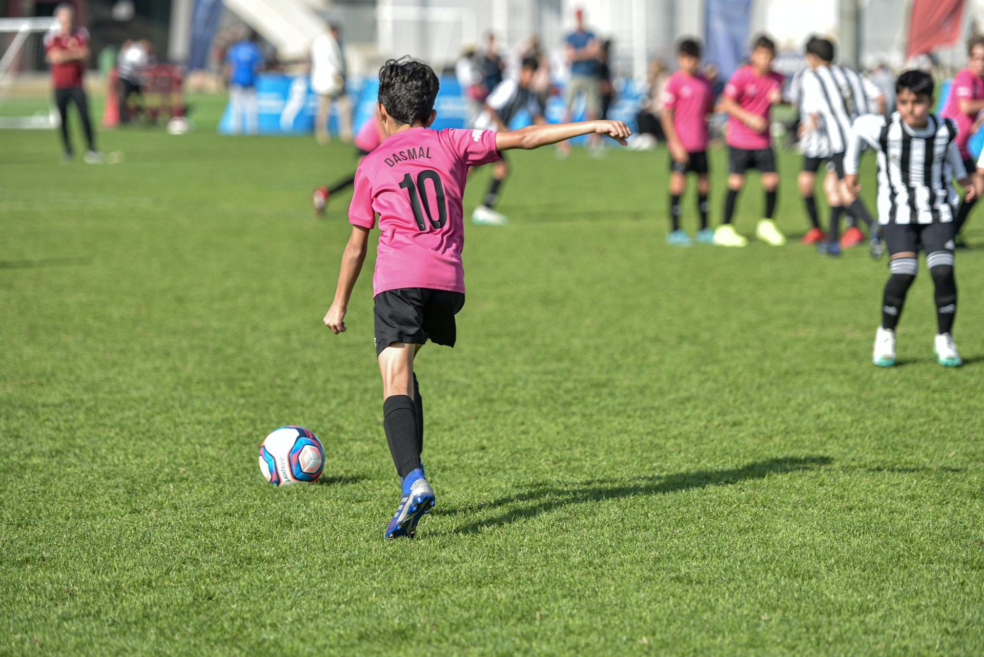 A soccer player in a pink and black uniform runs after the ball while their teammates stand on the sidelines, watching.