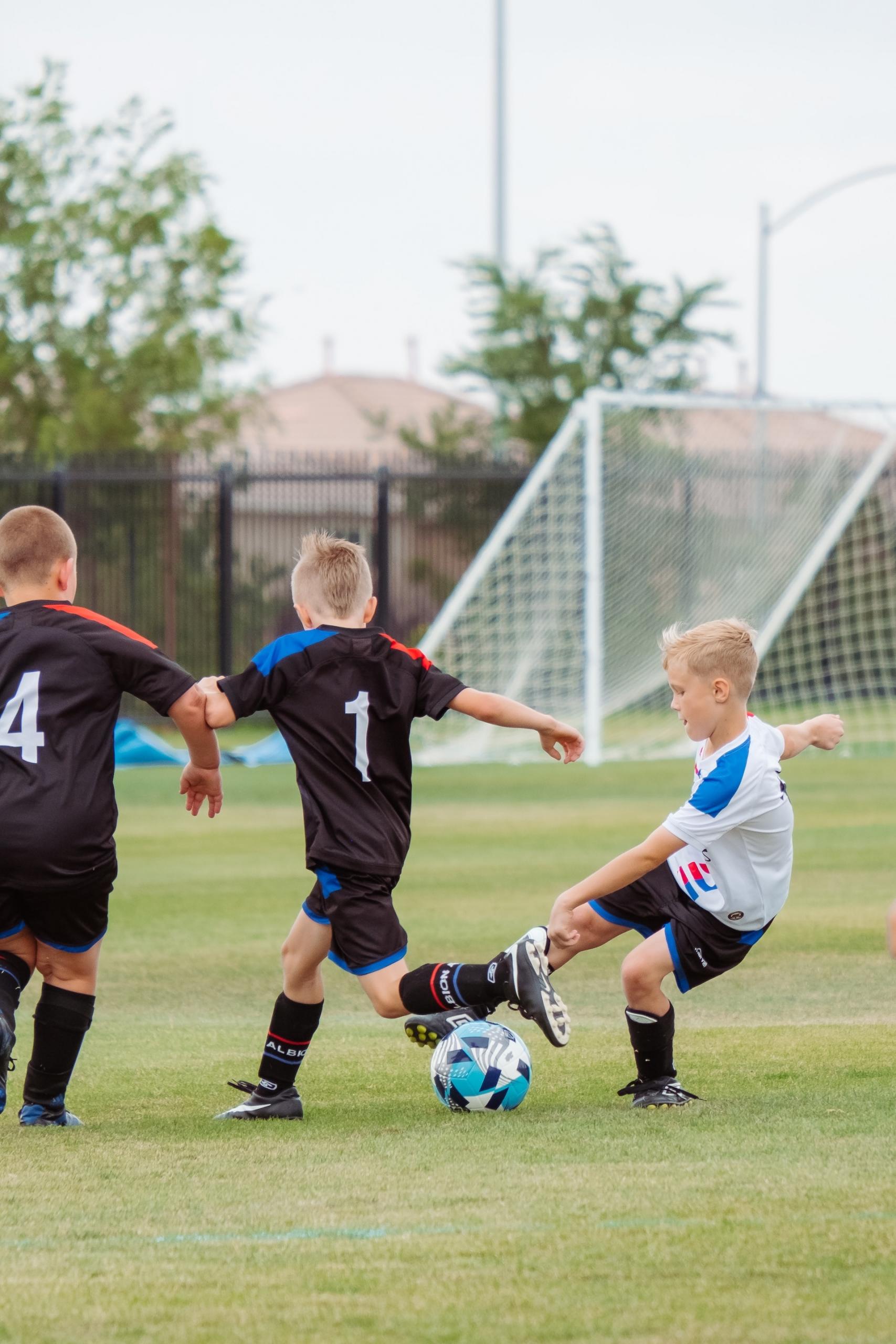 Two boys wearing black soccer uniforms with white numbers on their jerseys try to take the ball away from an opposing player who is wearing a white soccer jersey with light blue shoulder patches and black shorts.