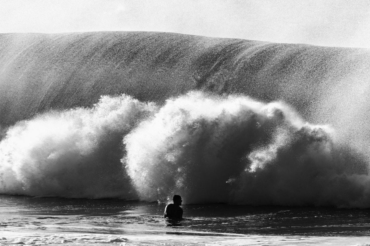 A greyscale image of a person standing chest deep in water in front of a cresting wave.