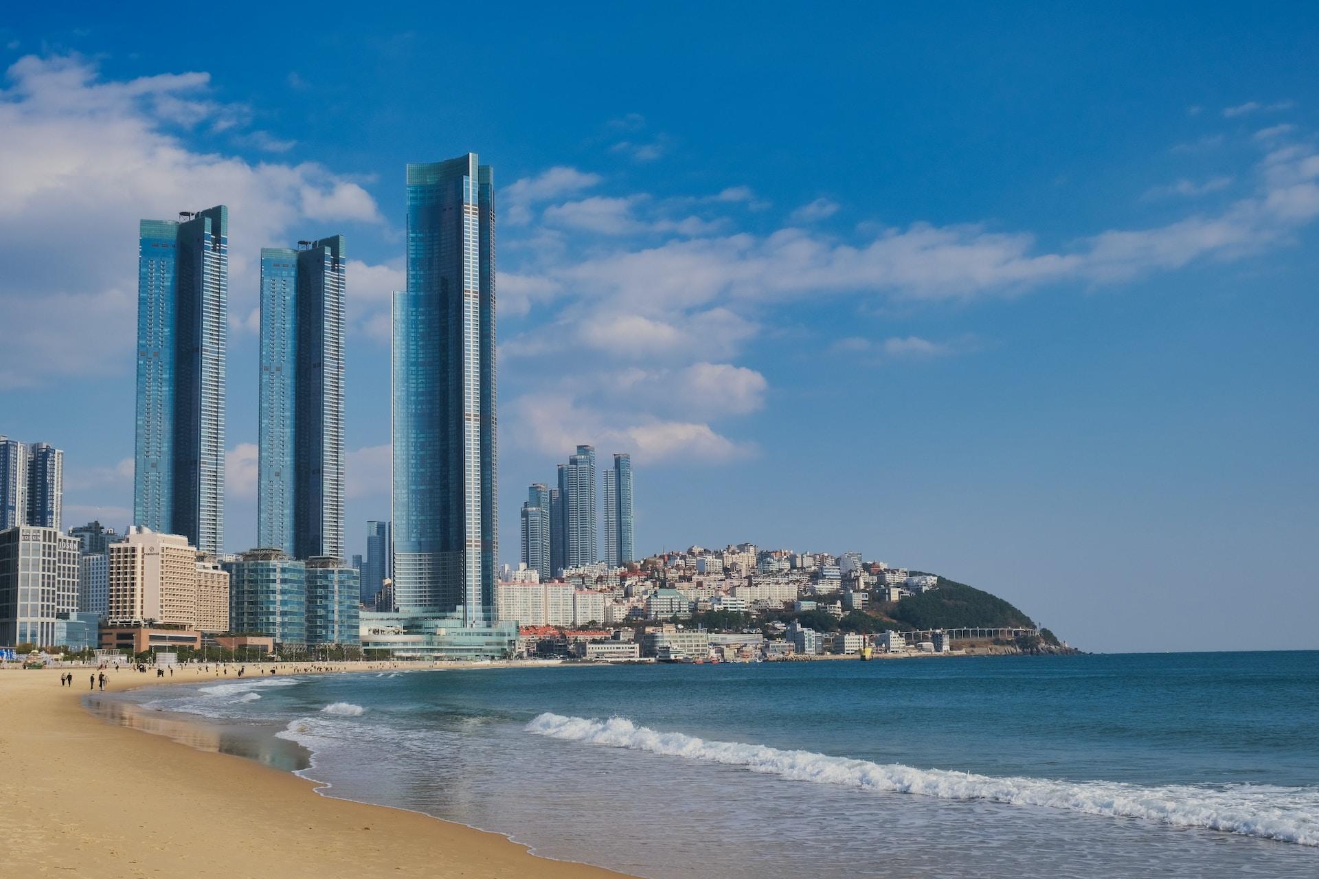 A view of Busan's silver skyline in the distance, with the beach and blue sea in the foreground under blue, lightly cloudy skies.