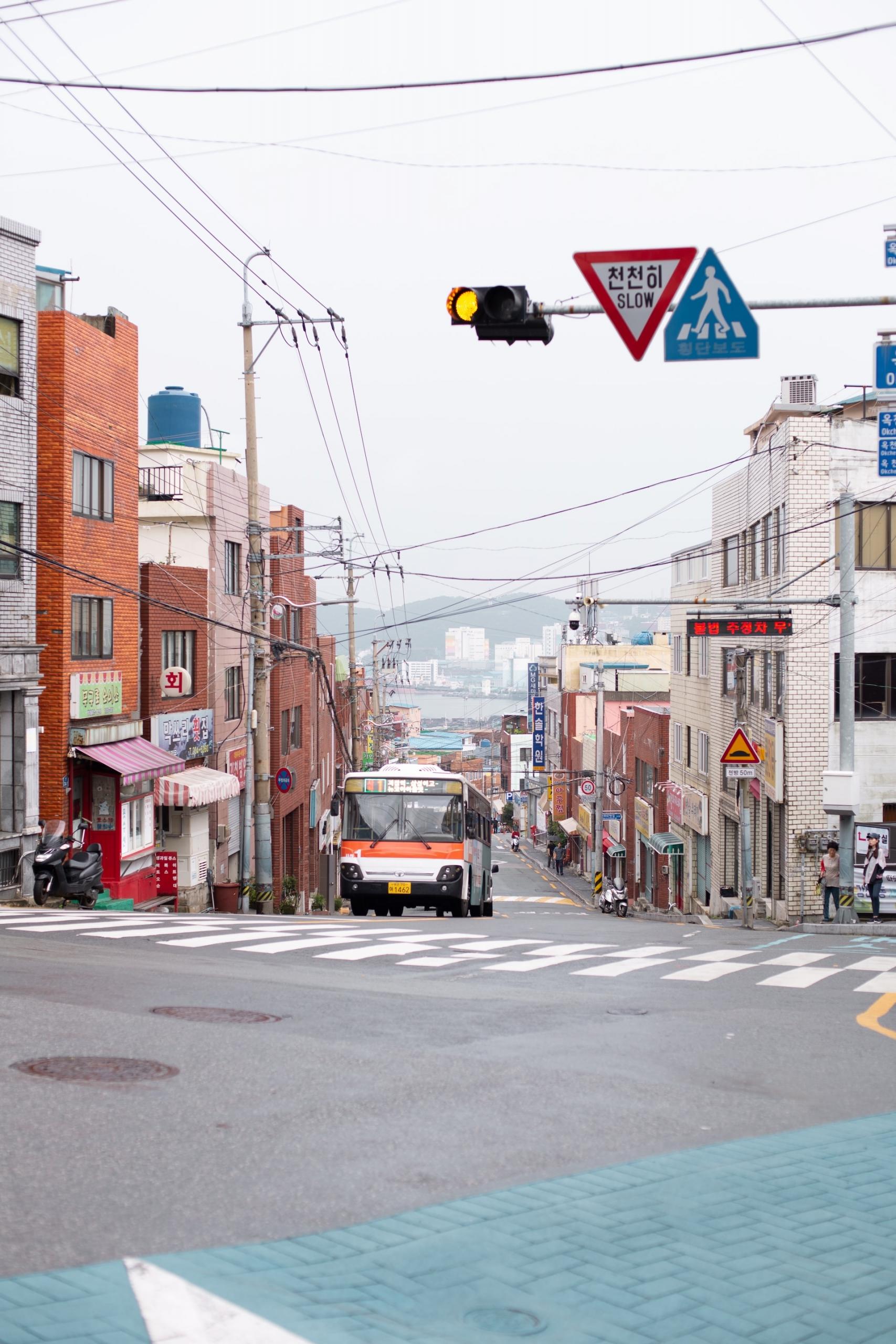 A street view of Busan with commercial establishments on either side of the road, with an orange and white bus trundling up the road, stopping just ahead of the zebra stripes.