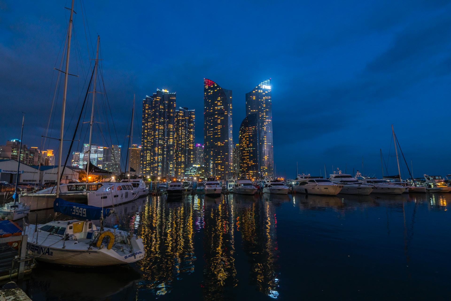 Busan's high-rise buildings seen from across the water at twilight, with boats docked in the foreground.