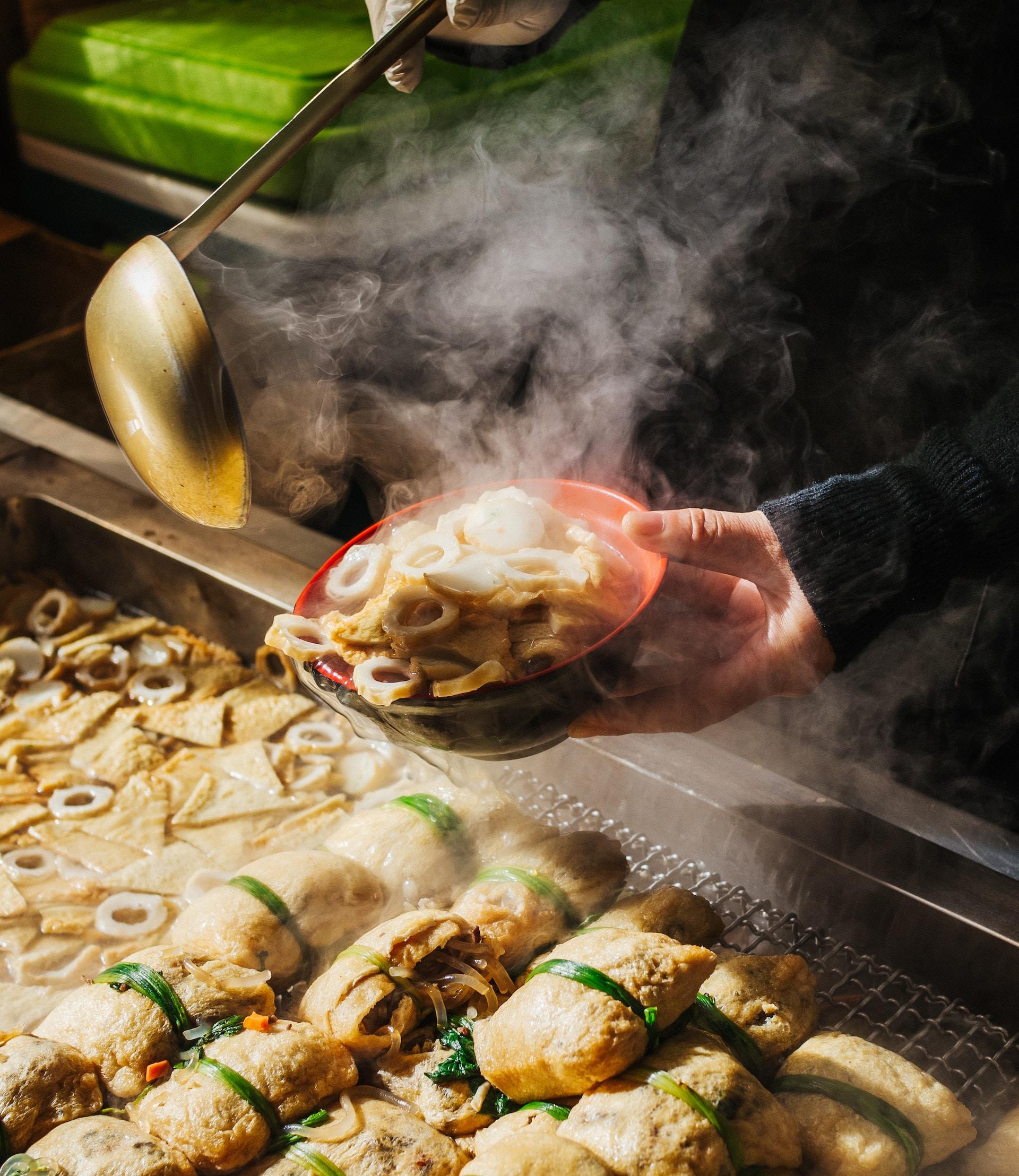 A food vendor in Korea ladles out a steaming portion of delight into a bowl with a red interior and black exterior.