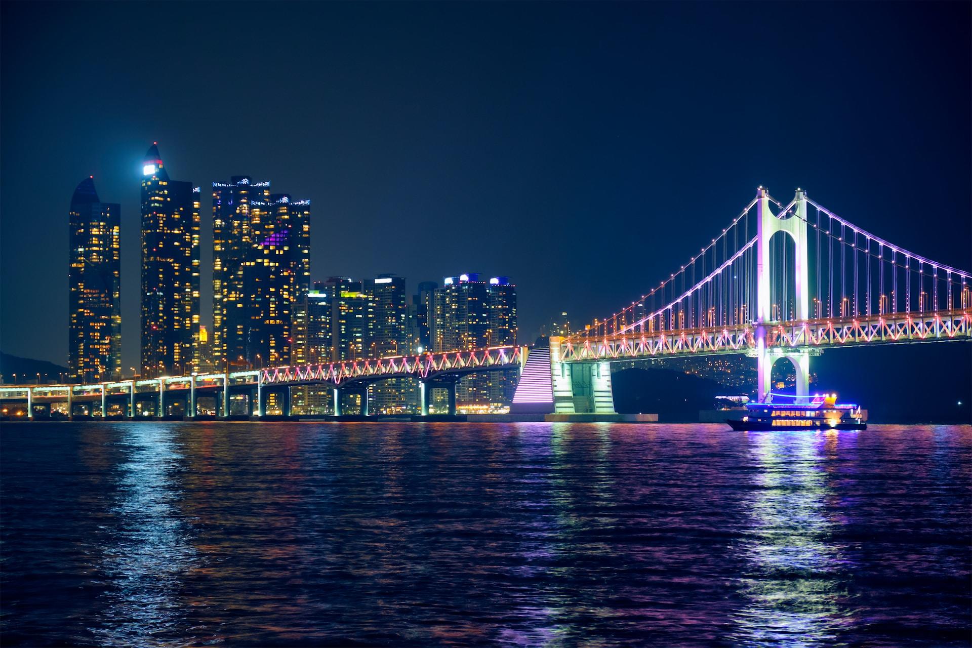 The Gwangan bridge at night, all lit up and reflecting in the water, with Busan's city lights forming a backdrop.