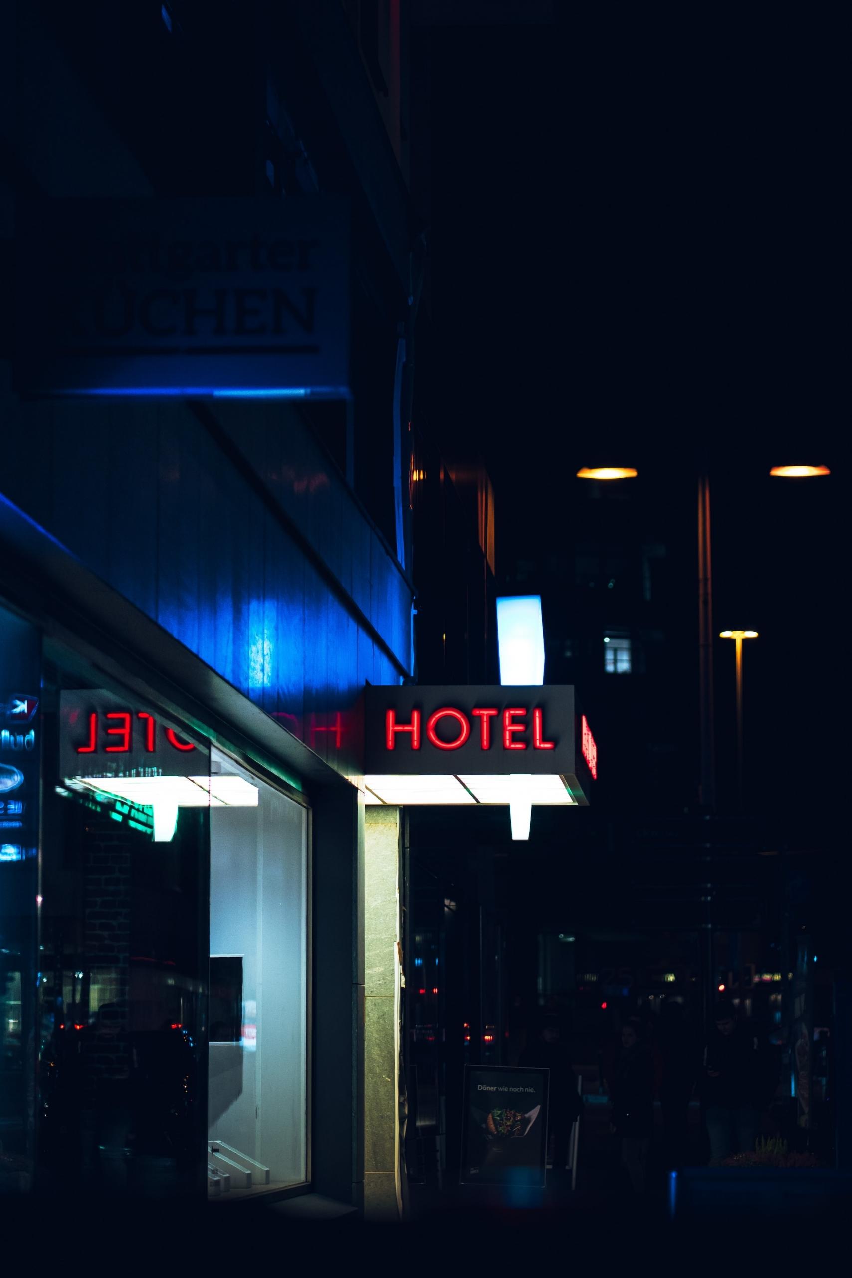 A red neon hotel sign along the awning over the facility's entrance contrasts against its electric blue facade in the night.