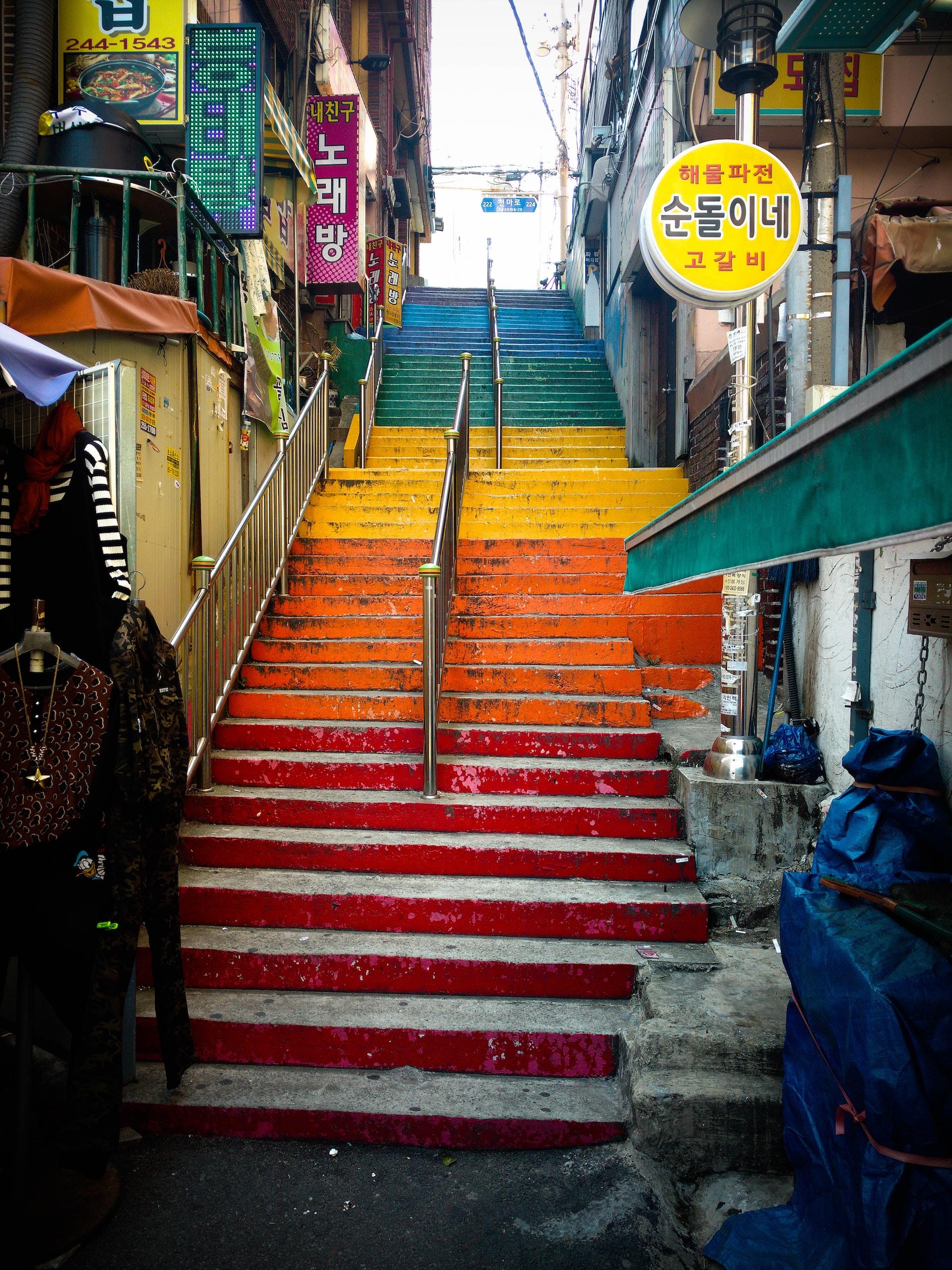 Busan's iconic outdoor colourful staircase, wedged between two blocks of buildings, with colourful signs overhanging the stairs and a handrail dividing it down the middle.