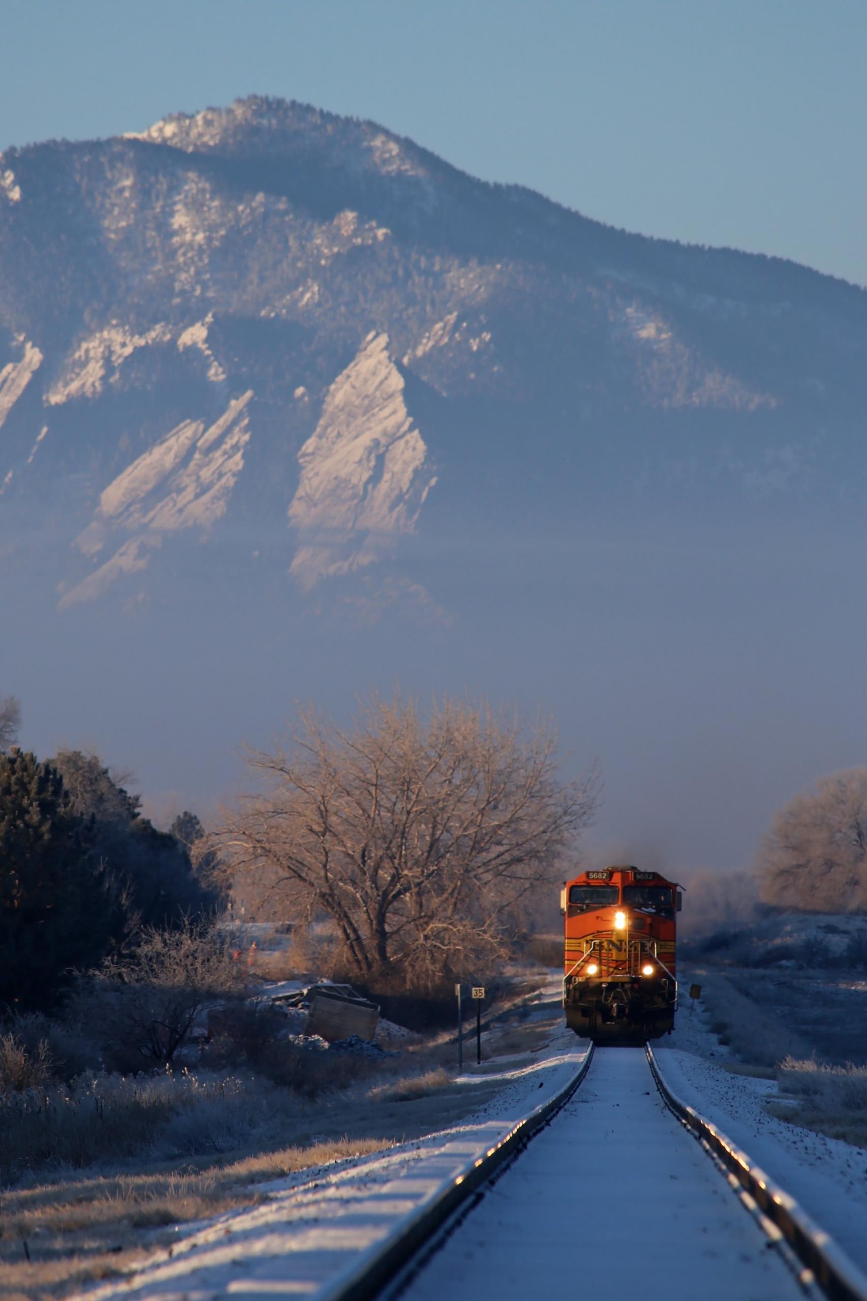 An orange train with its three lights on travels along a snowy track with a mountain in the background. 