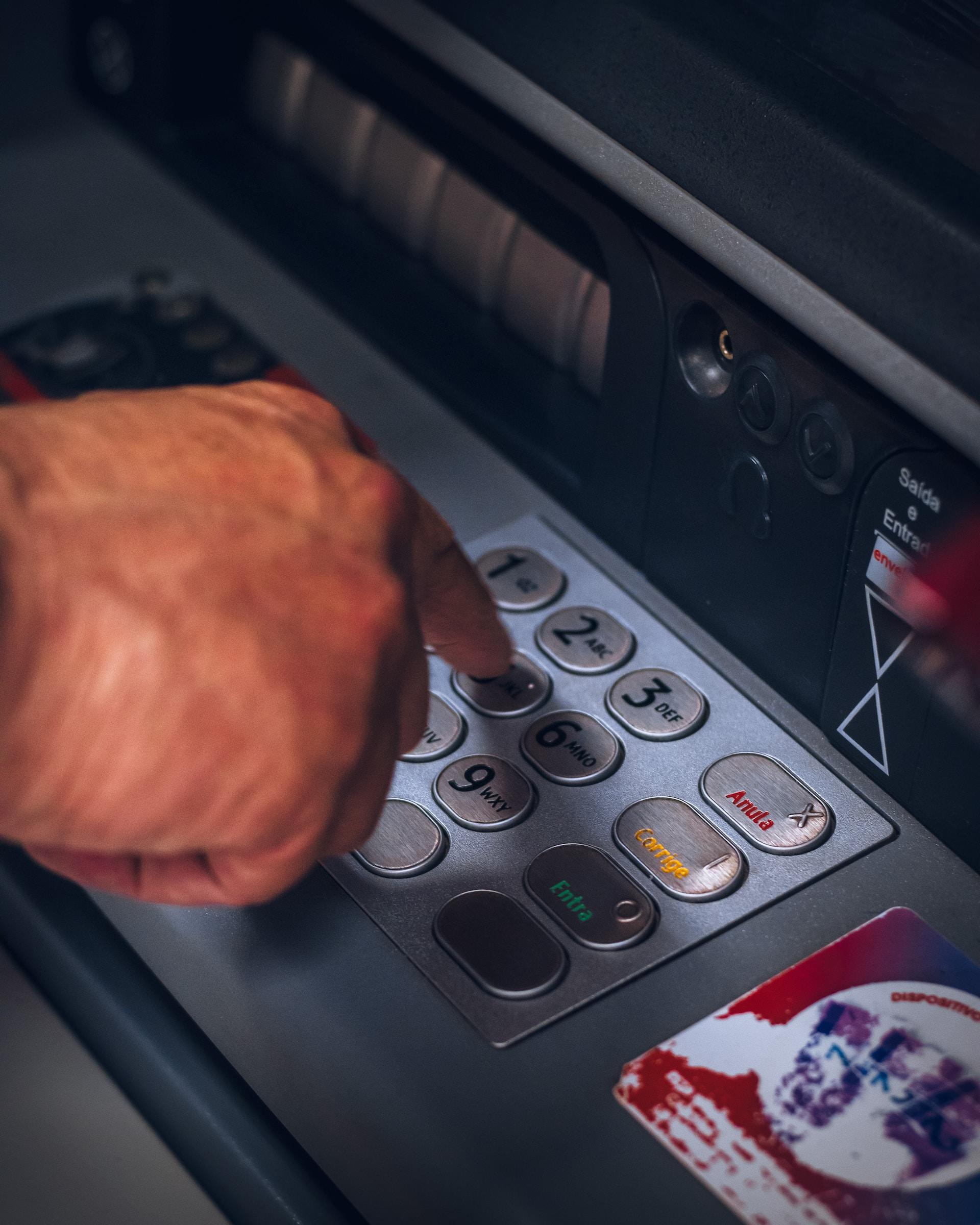 A person's right hand, index finger extended, hovers over a bank machine's keyboard, above the #5 key. 